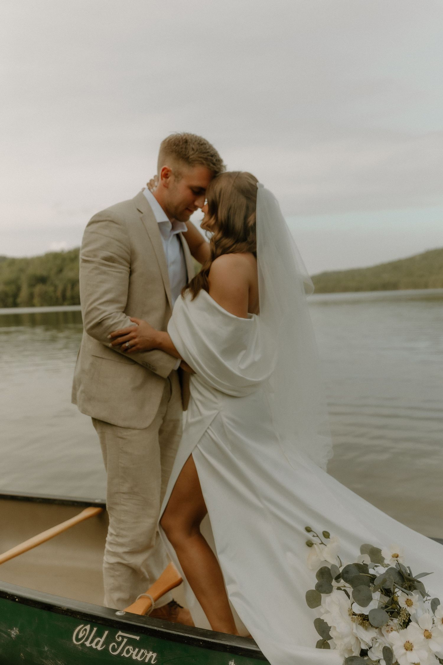 Newlyweds share an intimate moment while standing in an Old Town canoe on a lake.