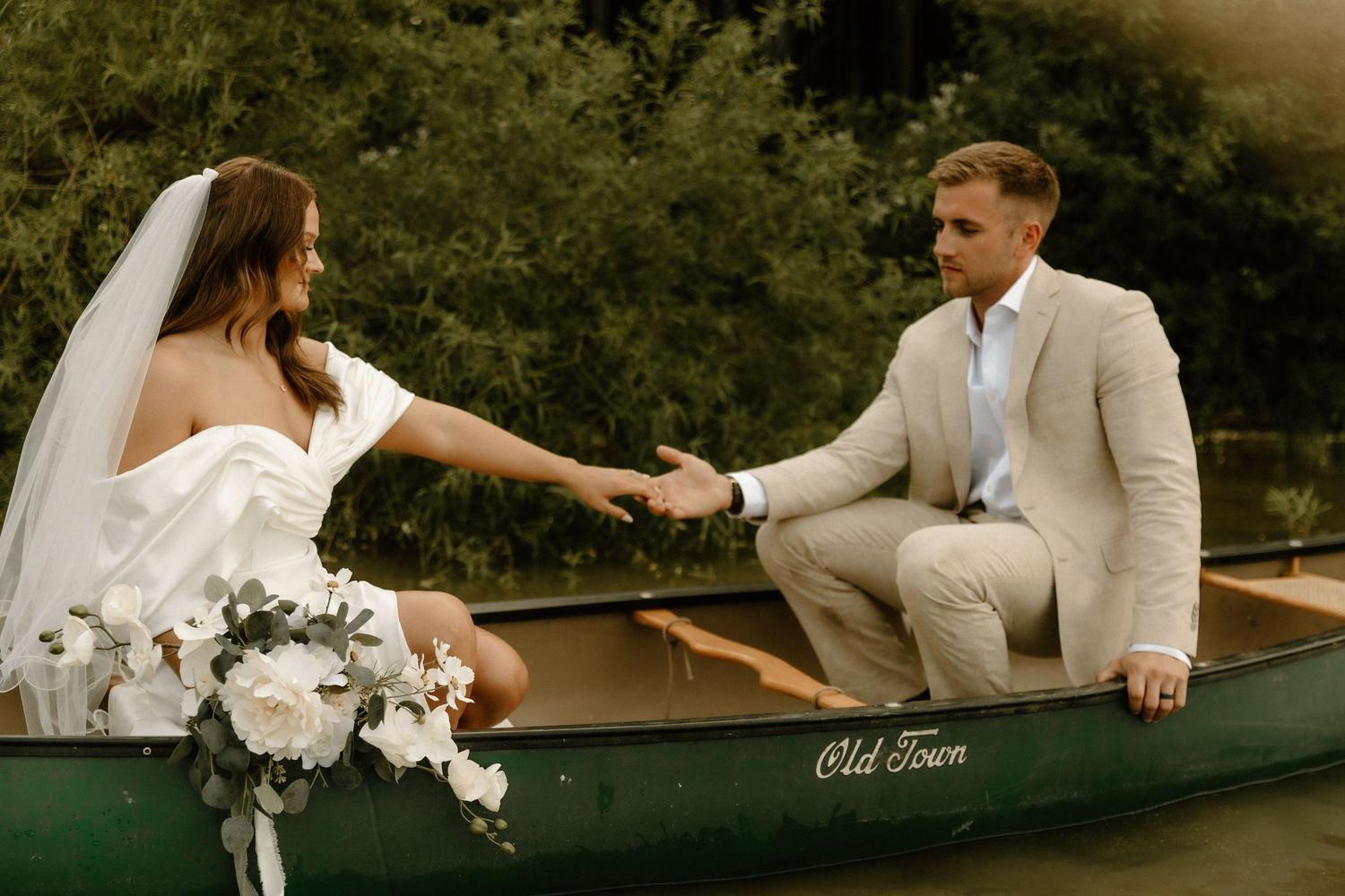 Bride and groom reaching for each other's hands while seated in a vintage canoe.