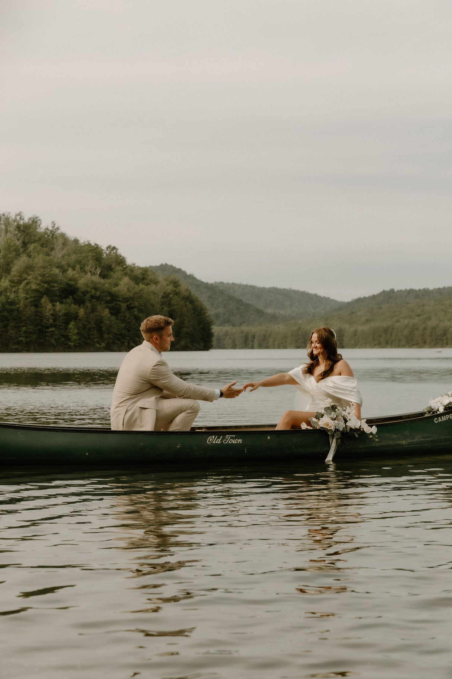 A couple in wedding attire sits facing each other in a canoe on a peaceful lake.
