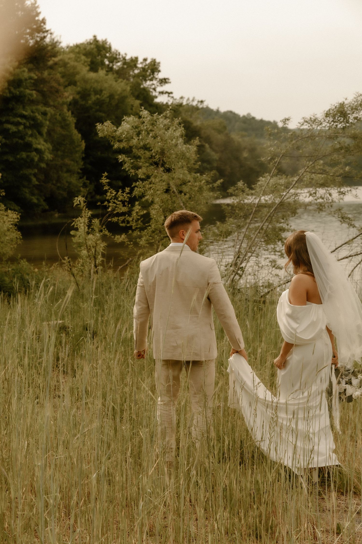 Couple in wedding attire walking through tall grass near a lakeside.