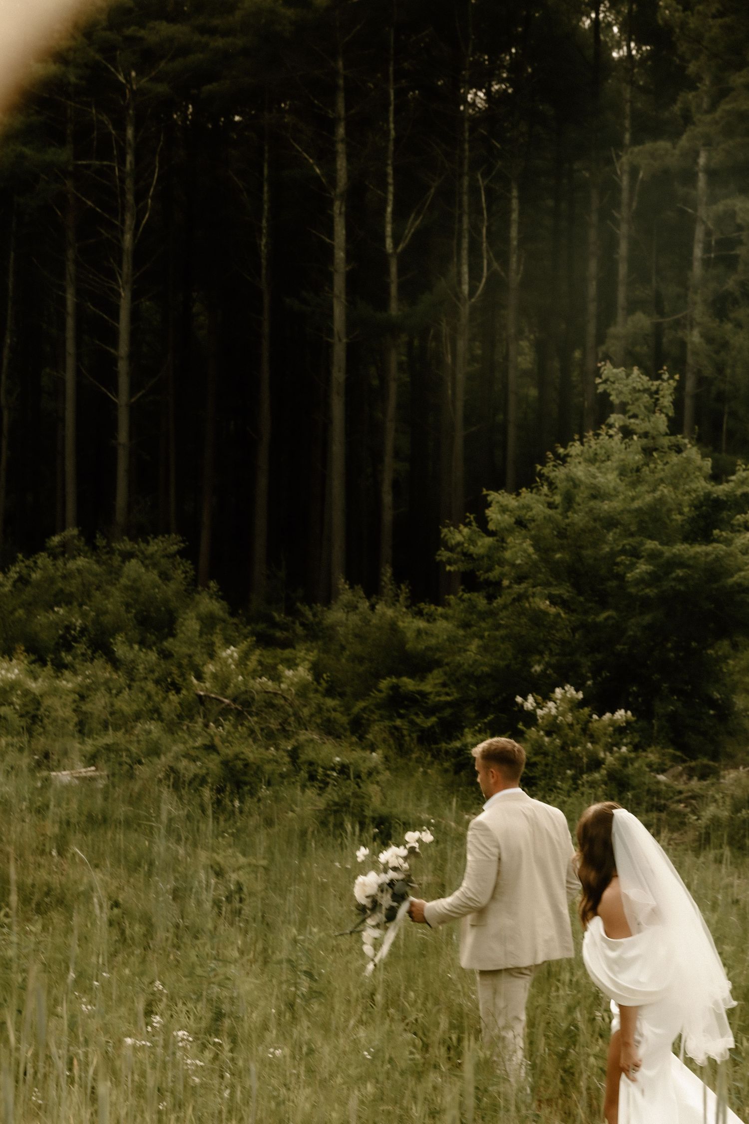 A wedding couple walks through tall grass near a dark forest holding flowers.