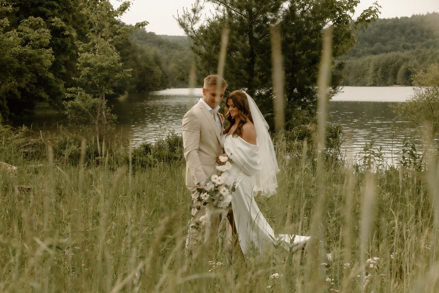 A couple in wedding attire poses among tall grass with a lake view behind them.