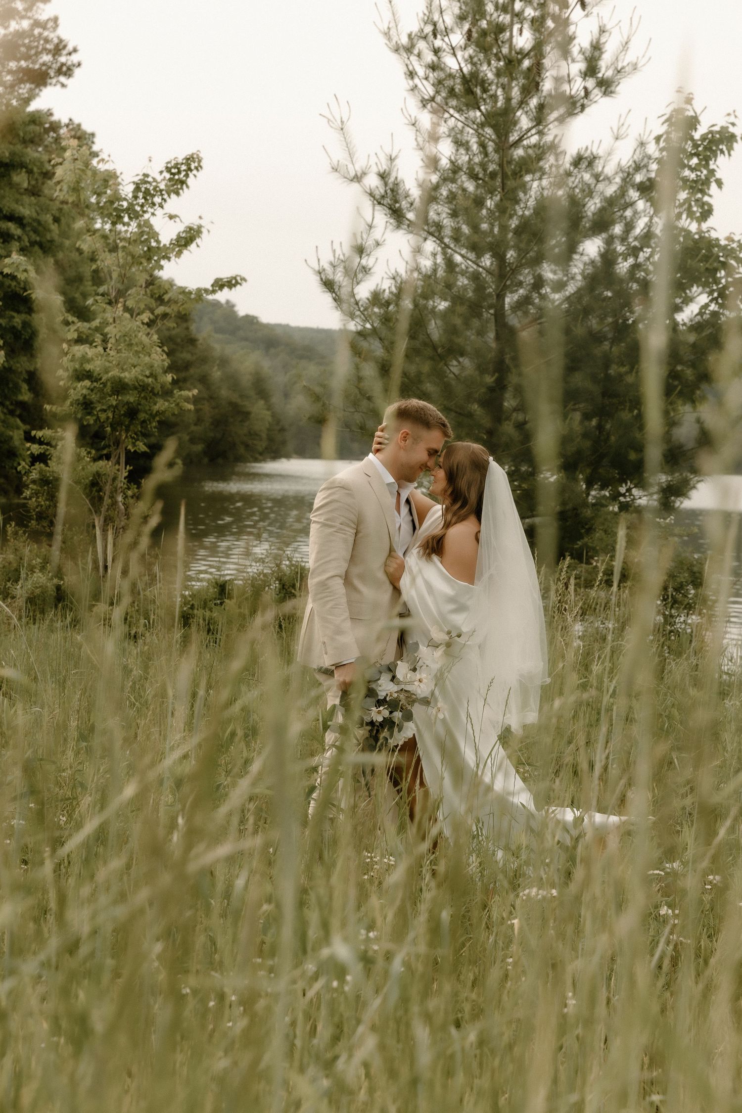 Newlyweds embrace in tall grass with a lake and mountains in the background.