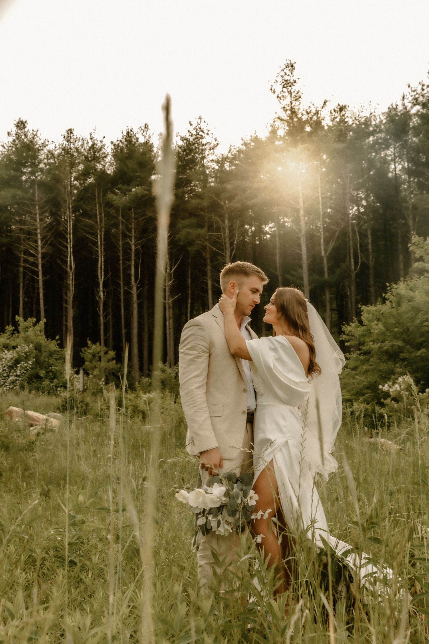 A romantic wedding photo in a sunlit forest meadow with tall pine trees in the background.