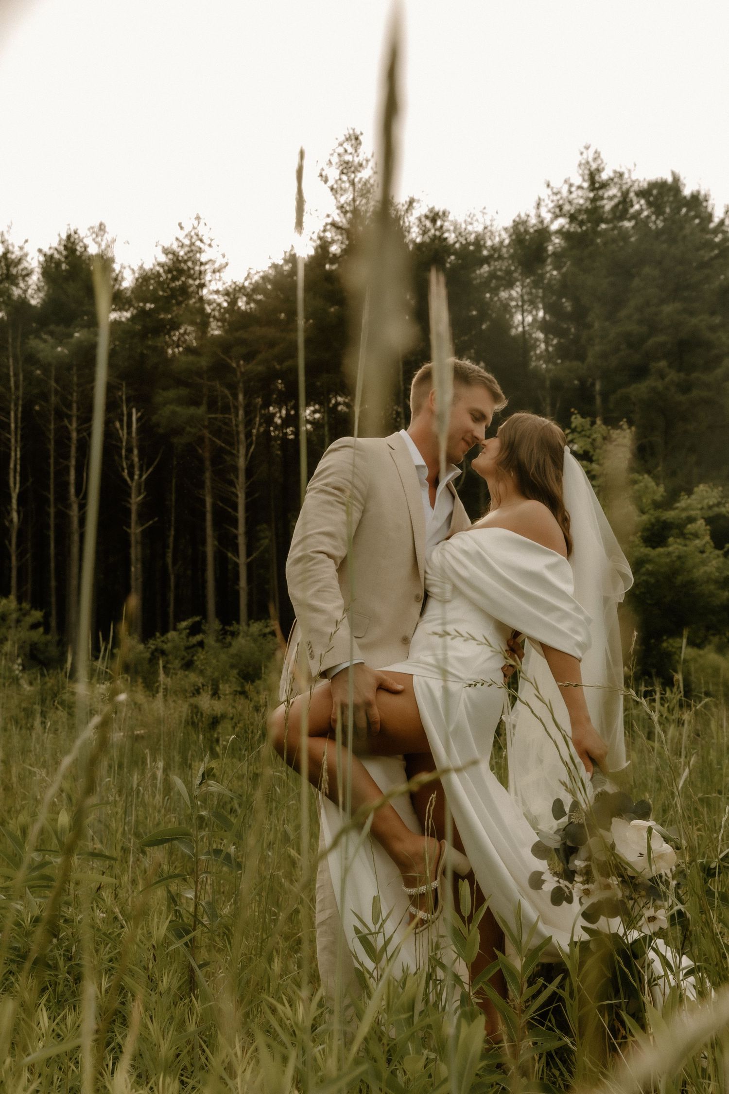 Artistic wedding photography through tall grass with pine forest backdrop.