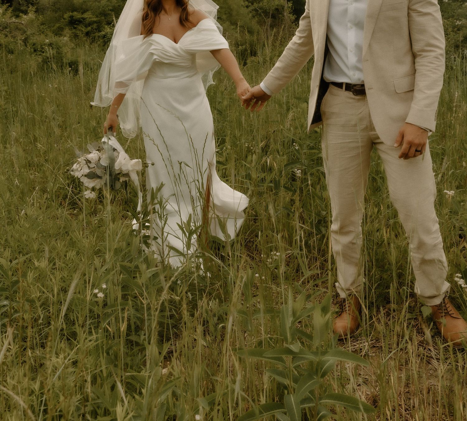 Elegant couple walking through wild grass in matching light-colored wedding attire.