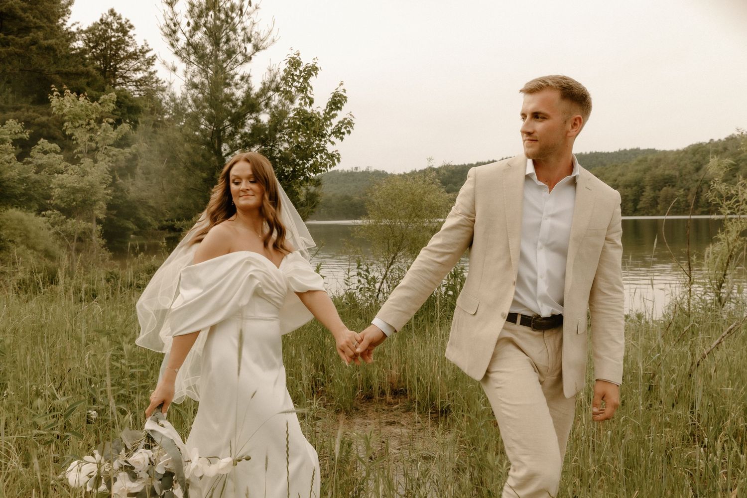 Newlyweds walking hand in hand through a meadow near a serene lake.