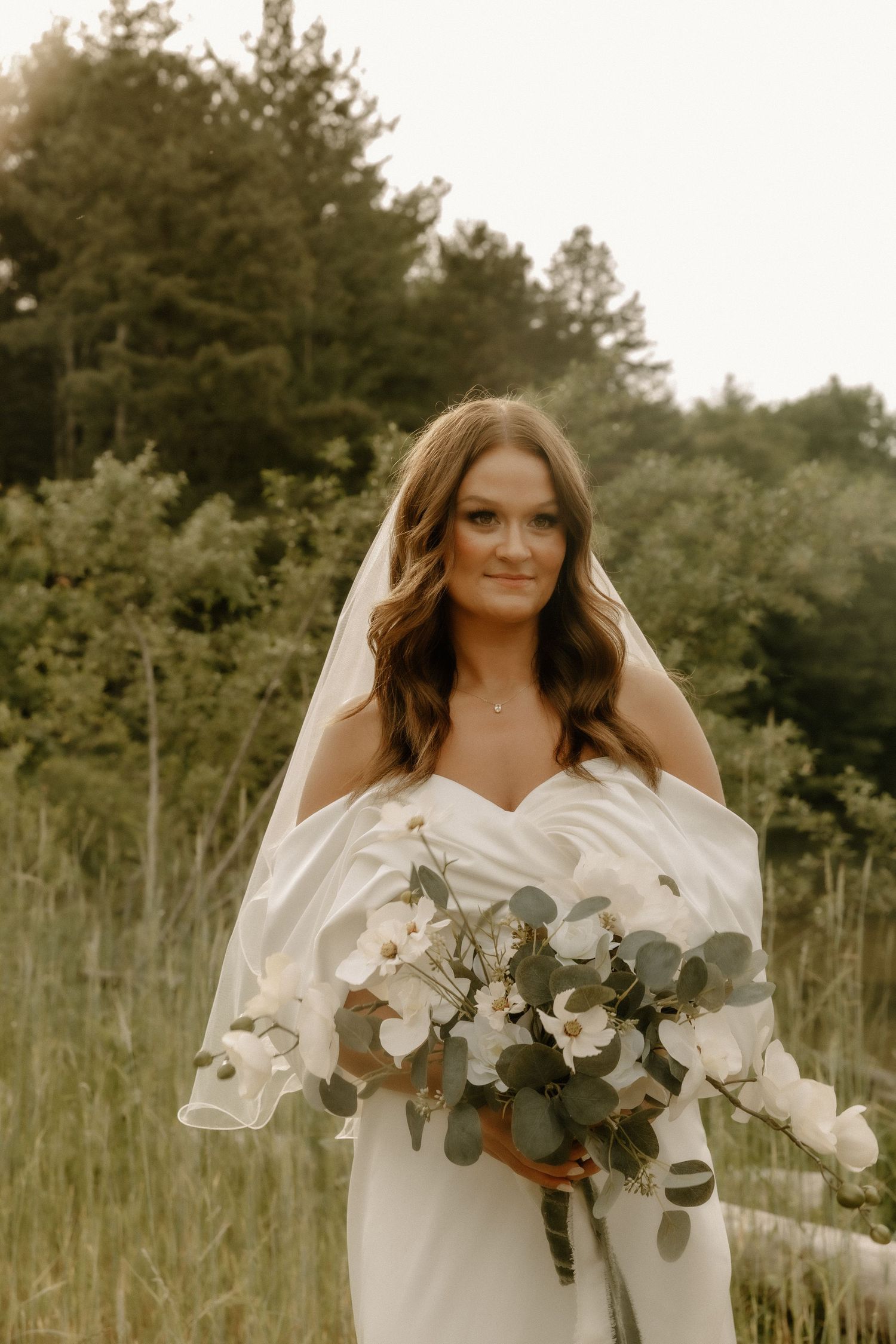 Bride holding a lush eucalyptus and white flower bouquet in an outdoor forest setting.