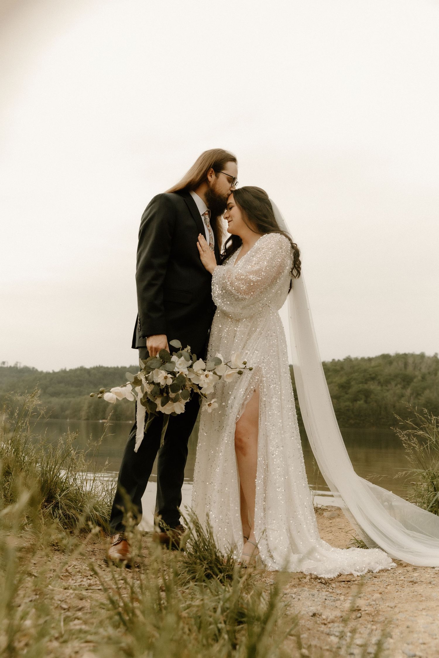 A romantic wedding portrait of a couple standing close together on a hilltop with the bride holding a floral bouquet.