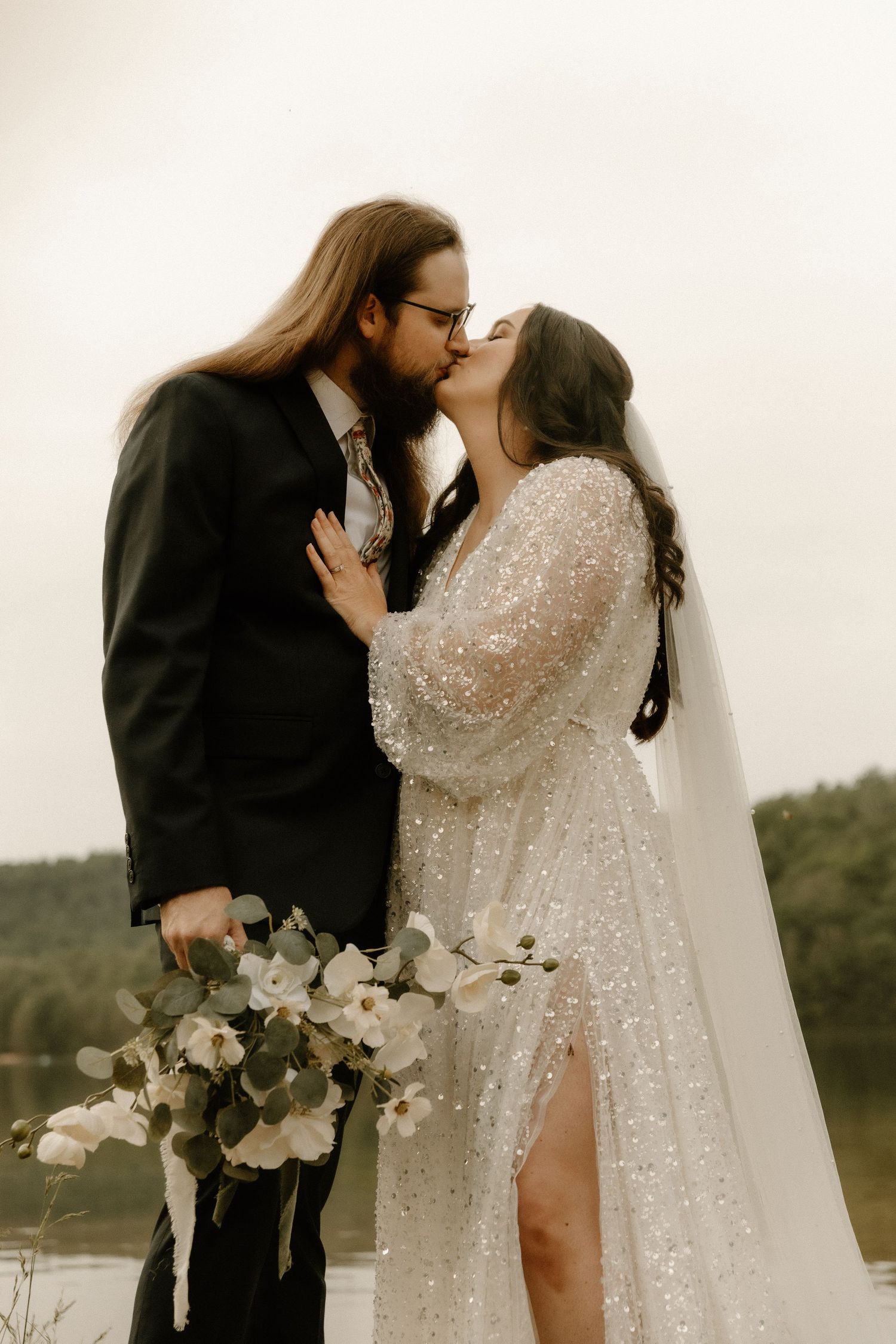 A couple in wedding attire shares a romantic kiss while holding a white floral bouquet.