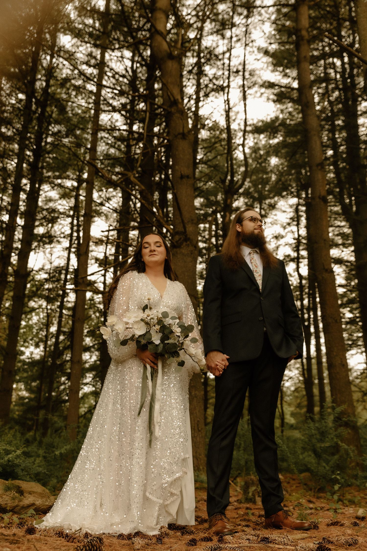 A wedding couple in black and sparkly white poses in front of tall pine trees.
