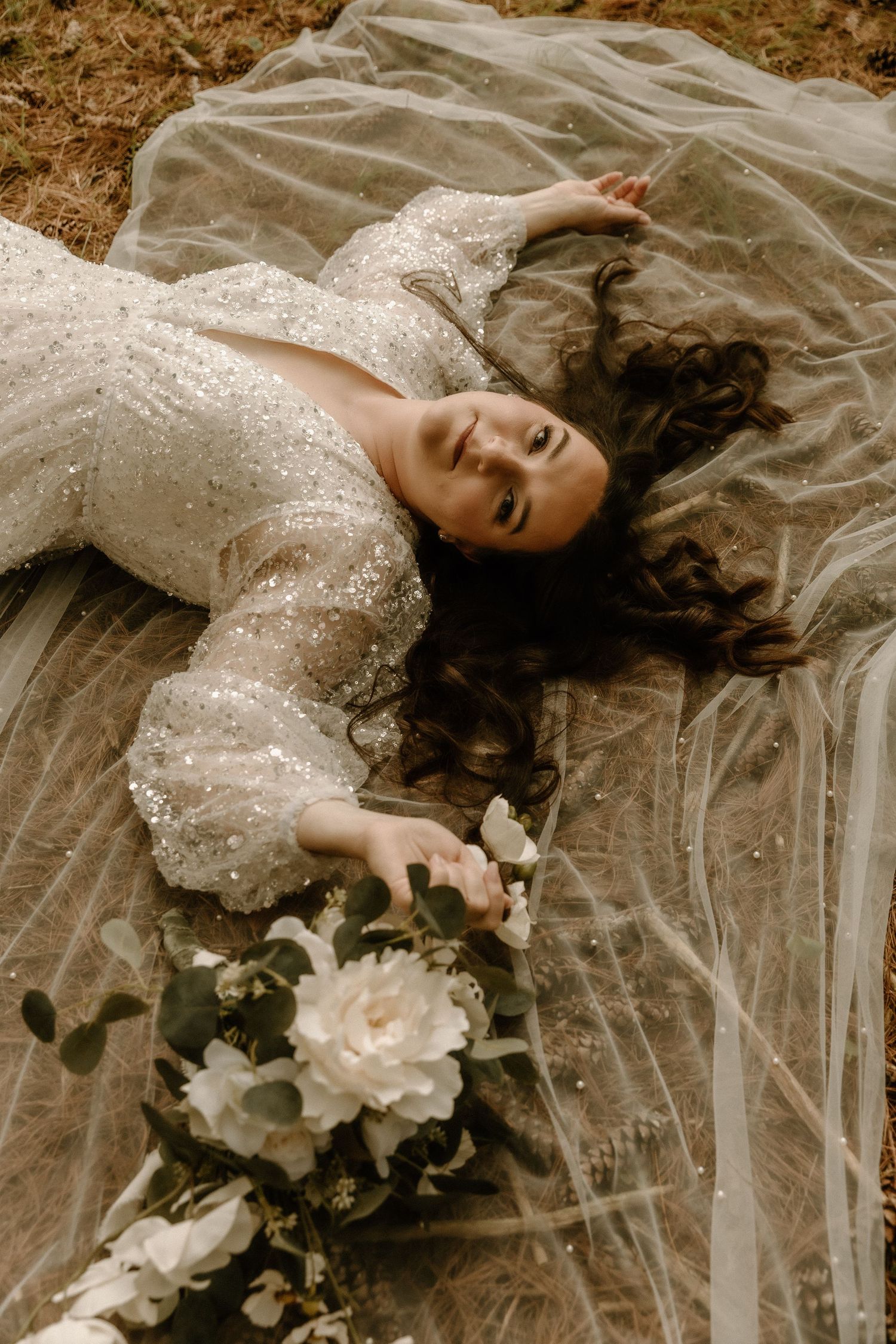 Bride in sequined gown lying on tulle veil with white flowers and flowing dark hair.