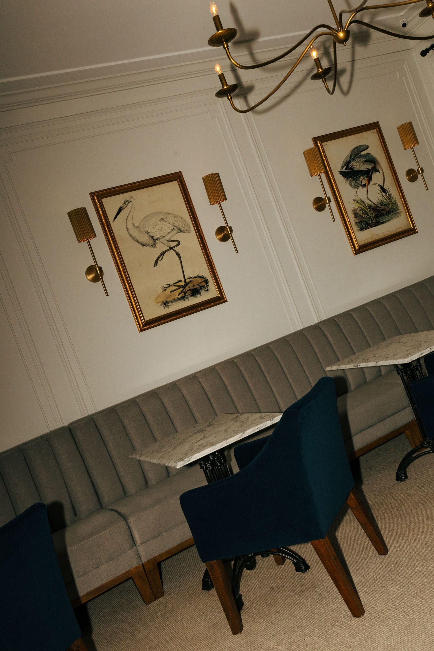 Vintage restaurant interior with blue velvet chairs, bench seating and framed bird artwork on white walls.