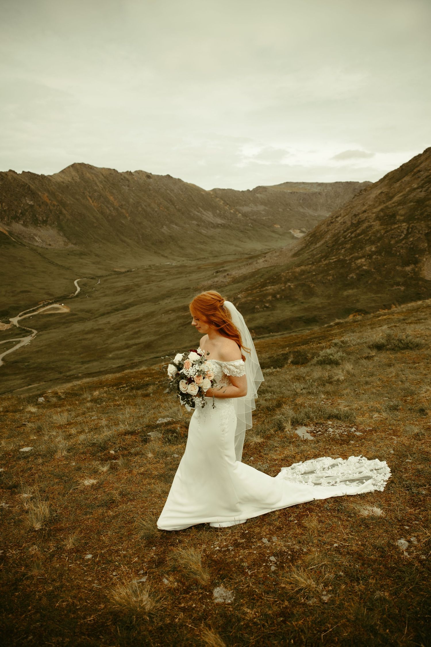 A bride in a flowing white gown holds a bouquet while standing on a mountain ridge overlooking a winding valley.