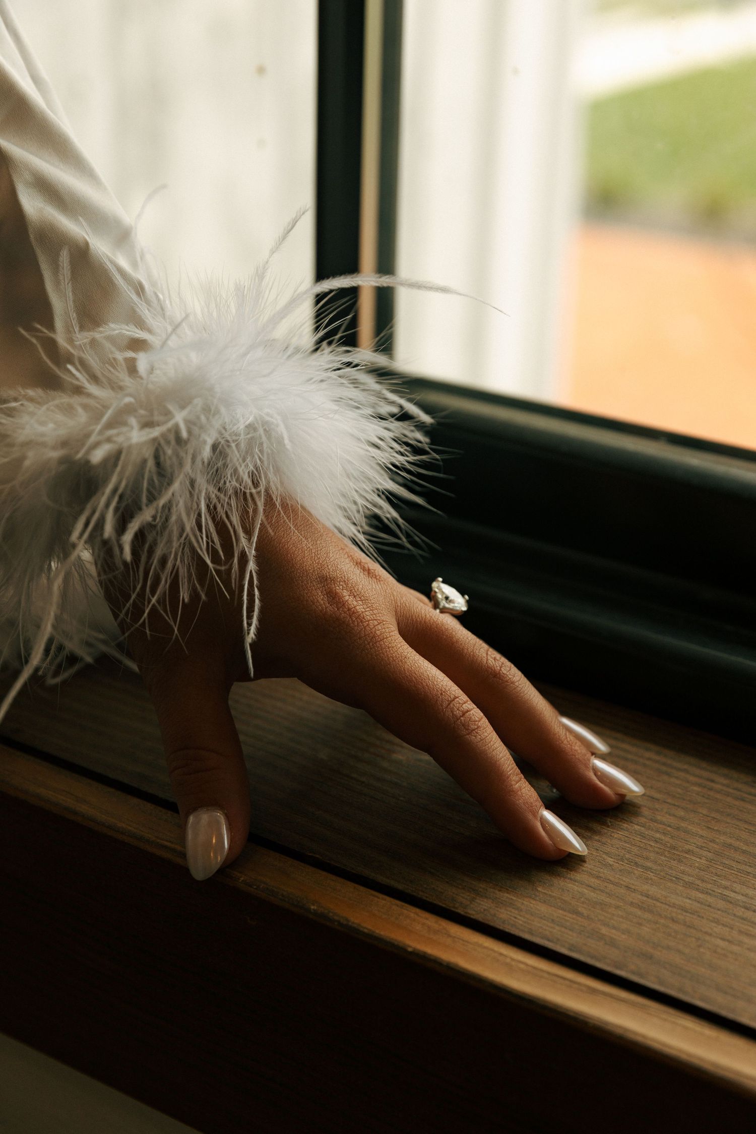 A hand wearing an engagement ring rests on a windowsill with delicate white feather details.
