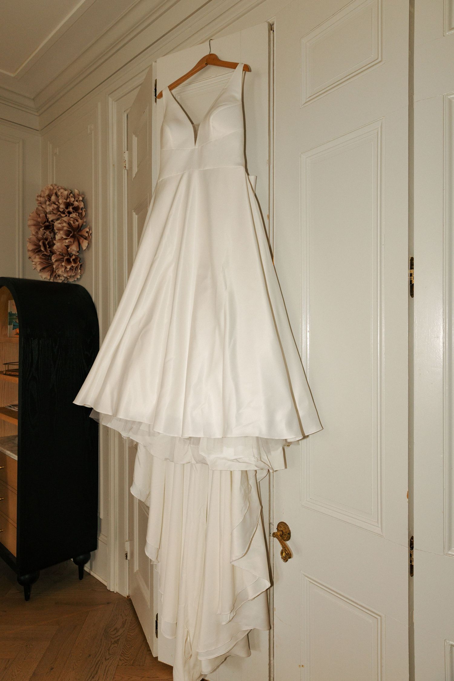 A white wedding gown with a sweetheart neckline elegantly hangs on a door next to a dark piece of furniture.