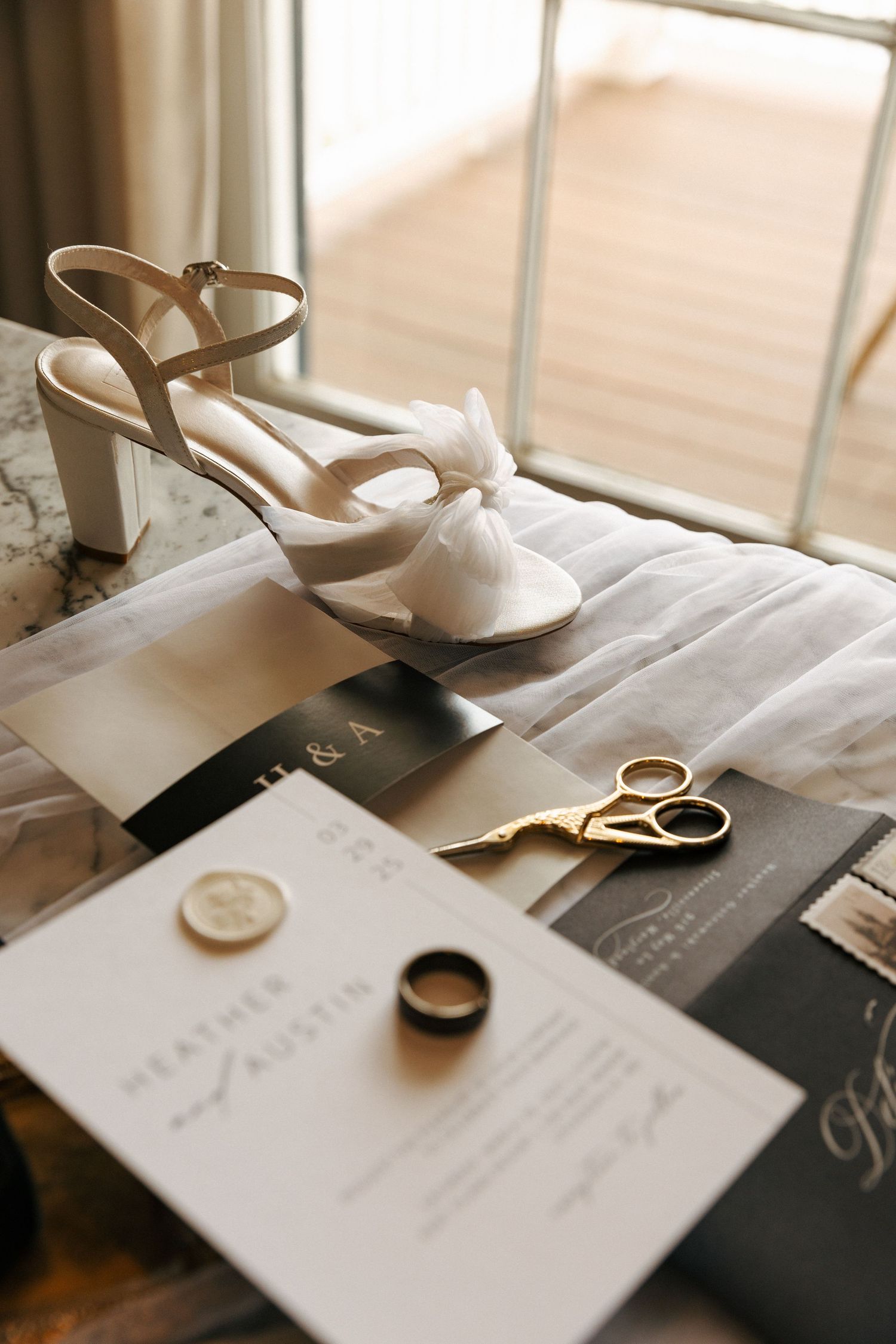 White bridal heels, wedding rings, and invitation details arranged on a surface near a window.