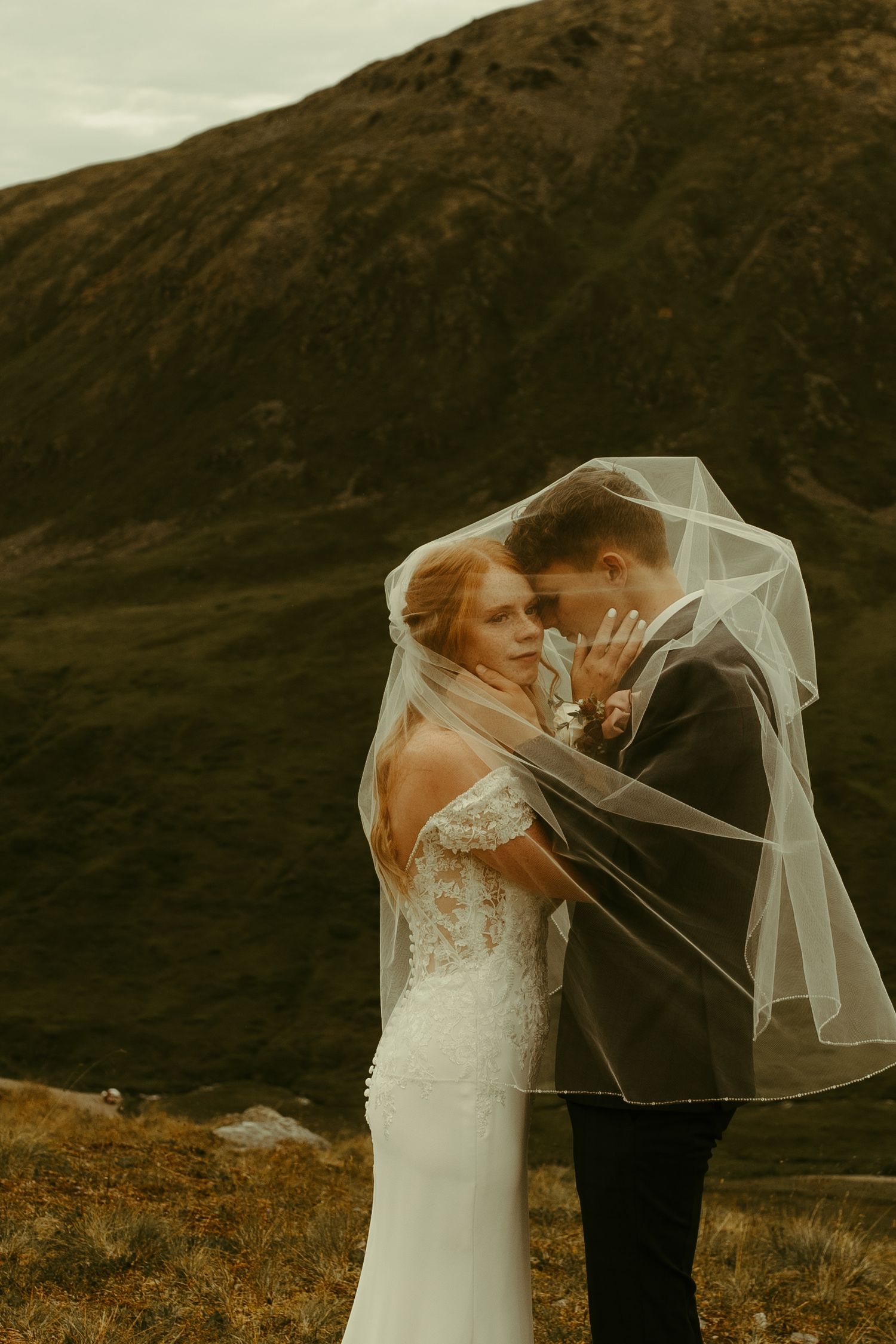 A romantic wedding couple shares an intimate moment beneath a flowing veil against a dramatic mountainous backdrop.