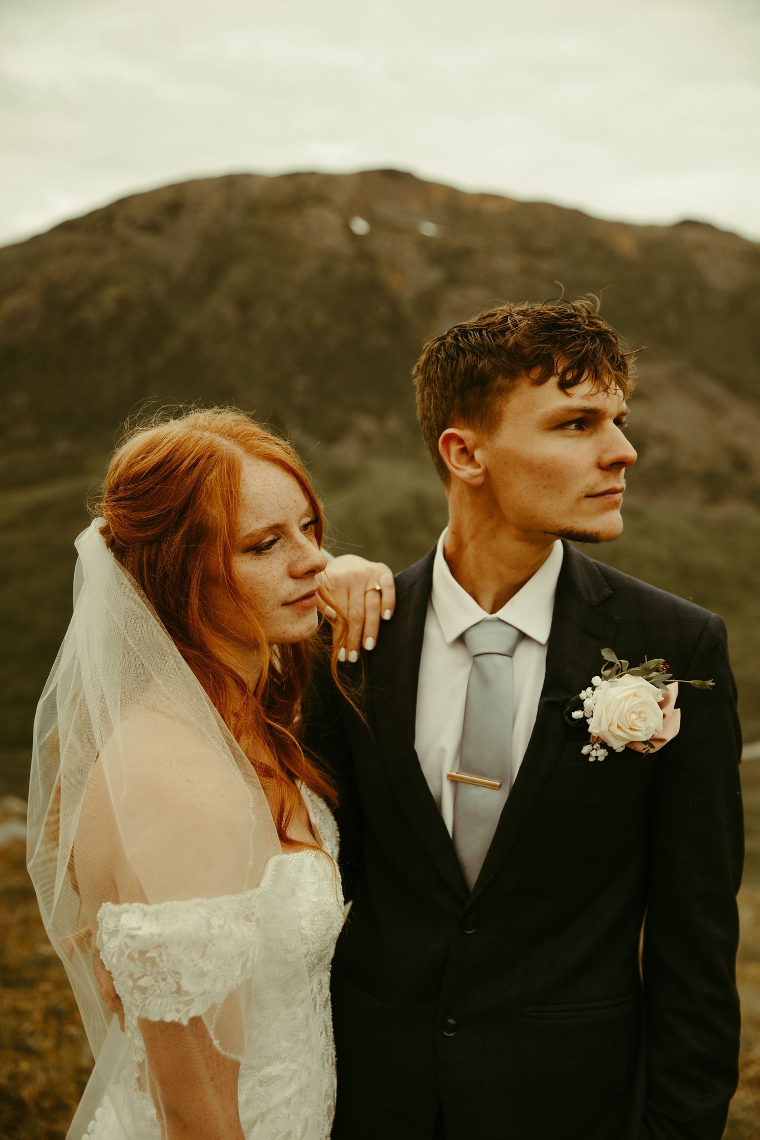 A bride and groom share an intimate moment outdoors in front of a moody mountain landscape at sunset.