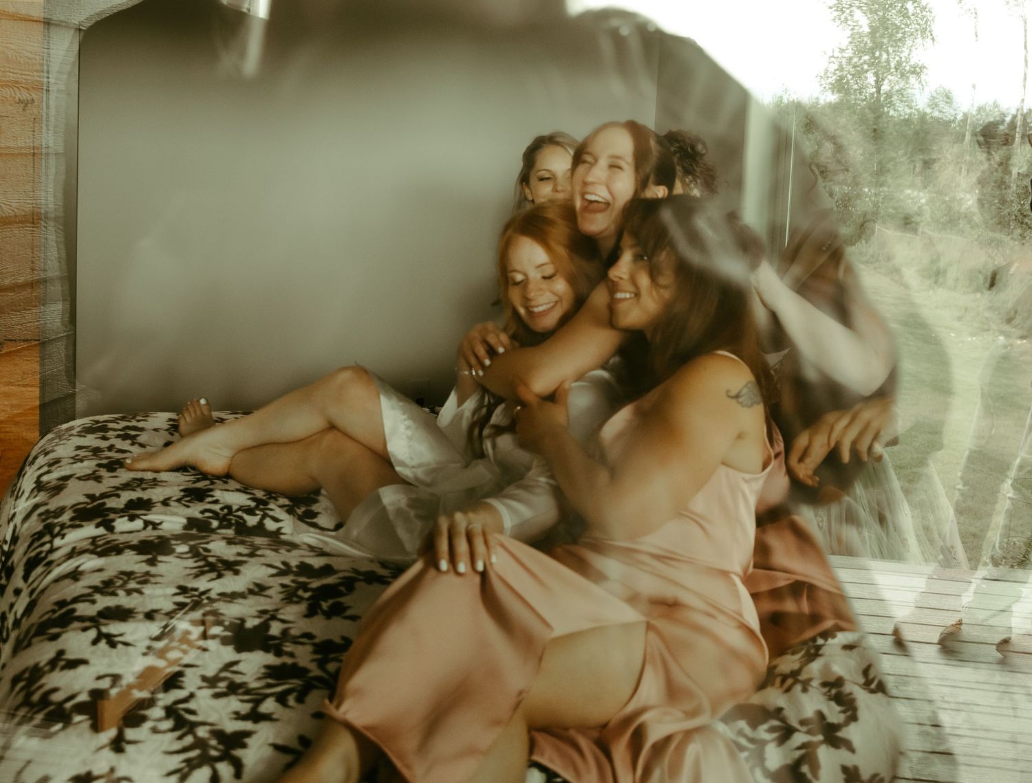 A dreamy sunlit bedroom scene with multiple people lounging together on a floral patterned bed.