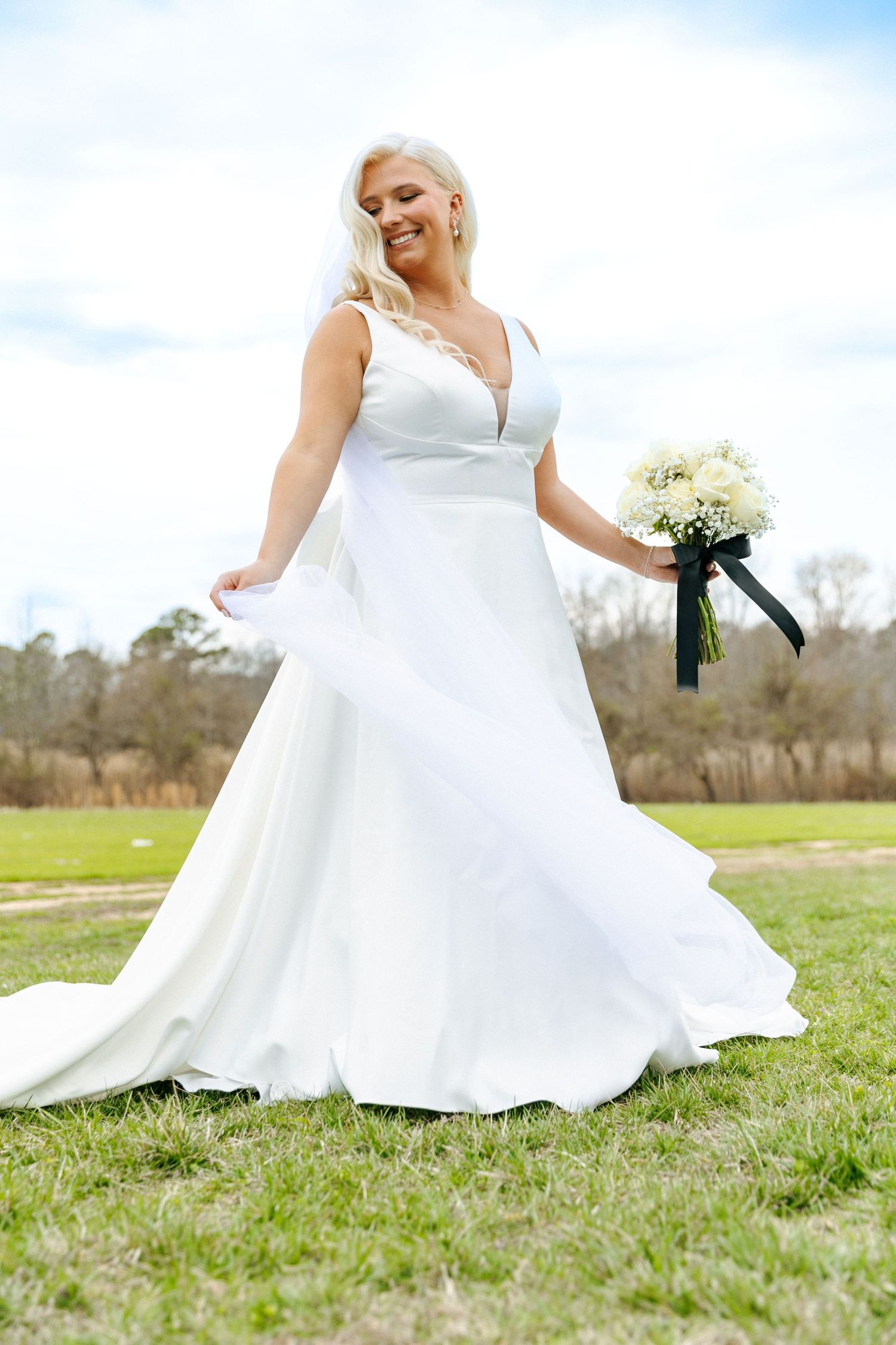 White wedding gown flowing in the breeze as bouquet is held against a natural outdoor backdrop.