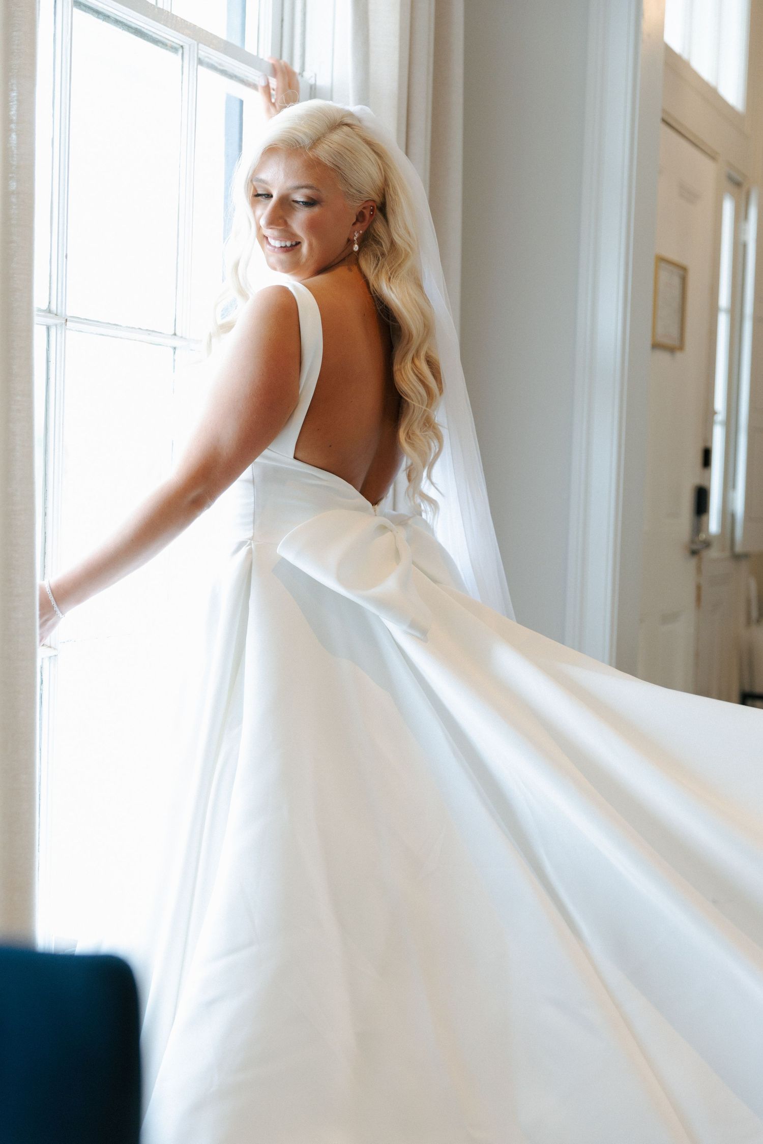 Elegant white wedding dress with an open back design and flowing train shown near a bright window.