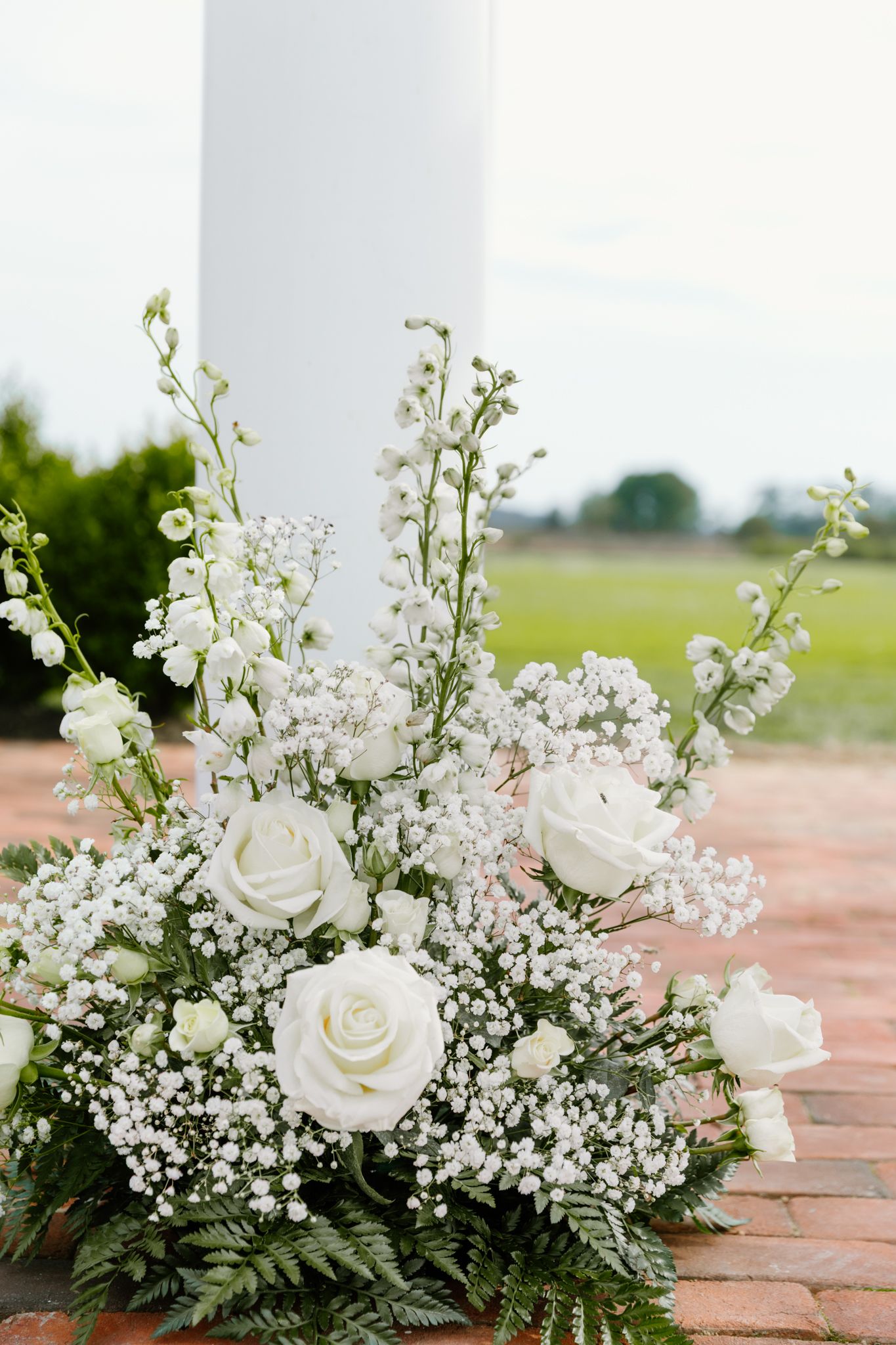 Elegant white rose and baby's breath floral arrangement displayed on brick patio with greenery and delphinium accents.