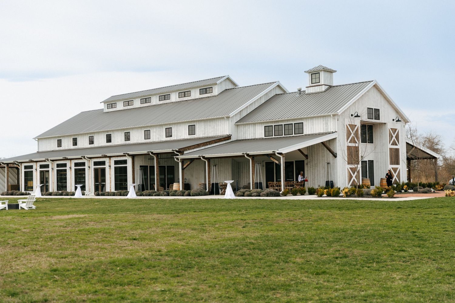 Modern white farmhouse style wedding venue with barn architecture and covered porches sits on a green lawn.