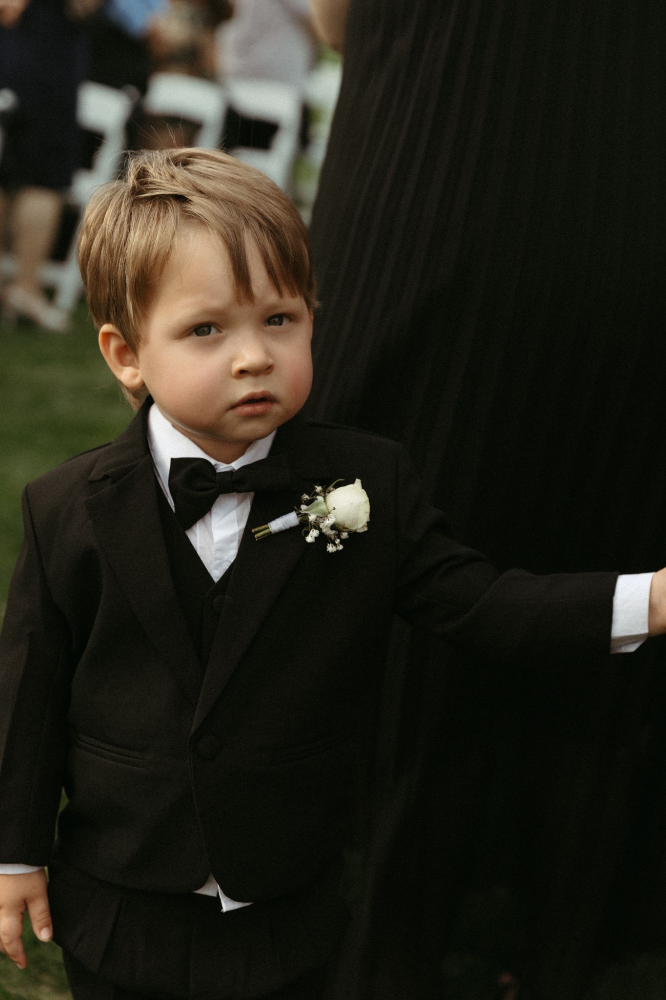 Young ring bearer in black tuxedo with bow tie and boutonniere stands at formal outdoor wedding ceremony.