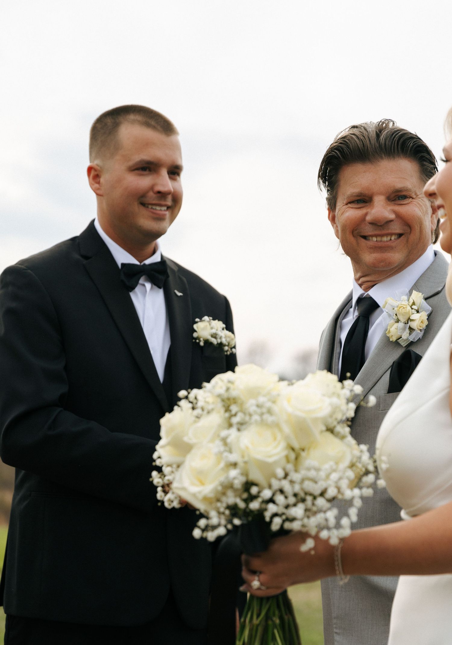 Wedding party members in formal attire stand together holding a beautiful white and baby's breath bouquet outdoors.