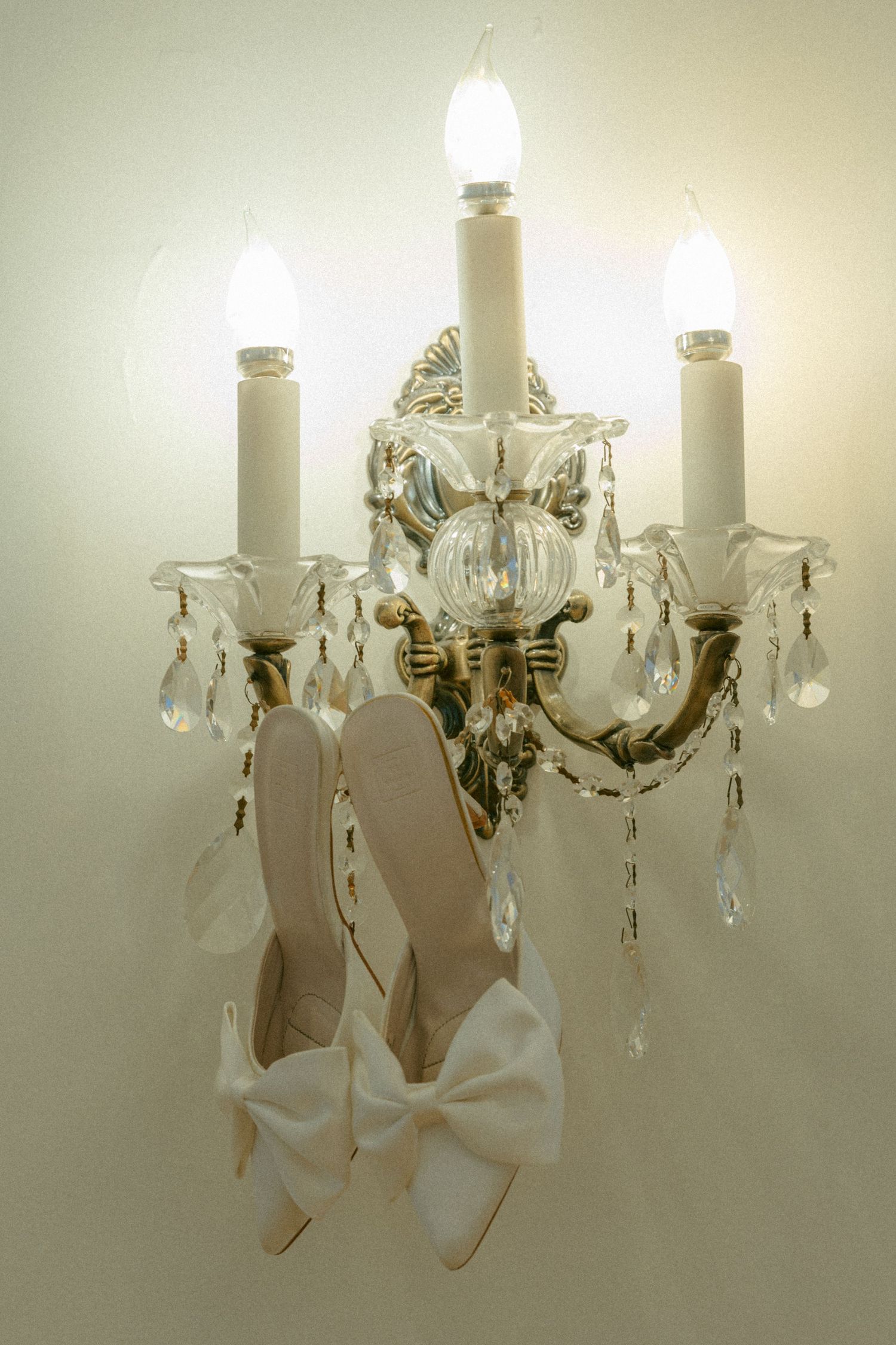 Elegant crystal chandelier wall sconce with hanging white bridal shoes featuring decorative bows.