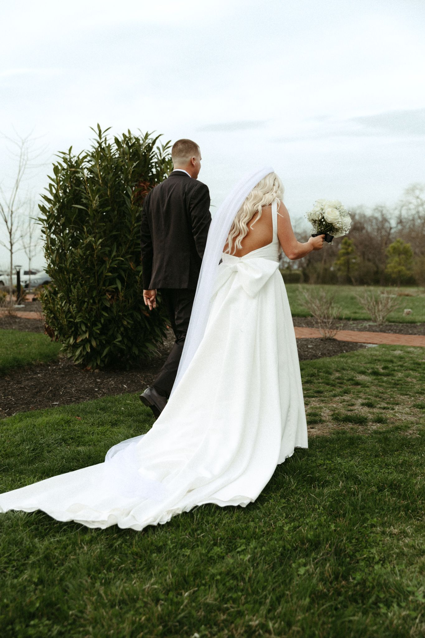Bride in flowing white gown with long train stands back-to-back with groom outdoors by a green bush on grassy lawn.