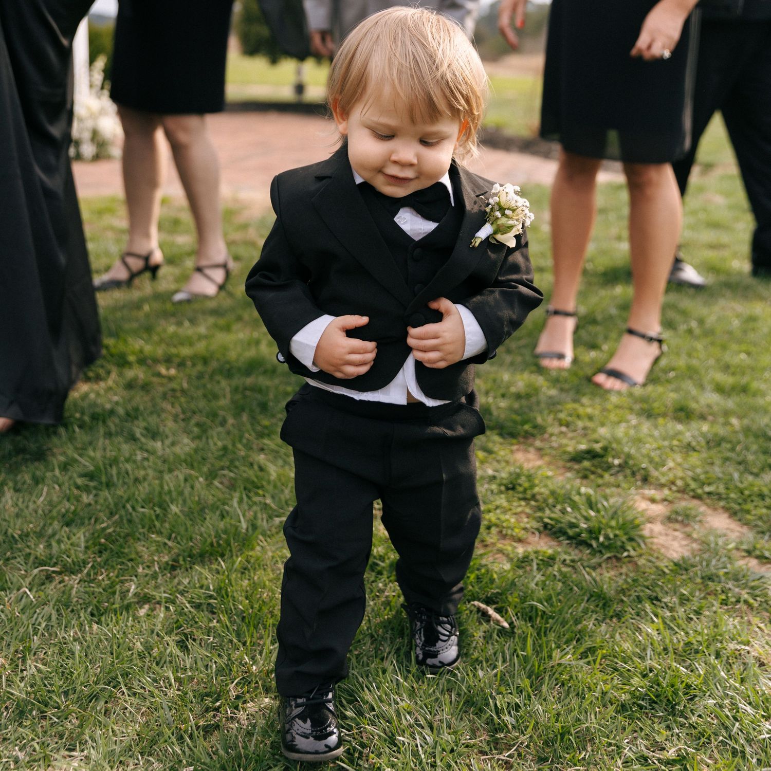 A young child wearing a formal black suit and boutonniere at an outdoor wedding stands on grass near other guests.