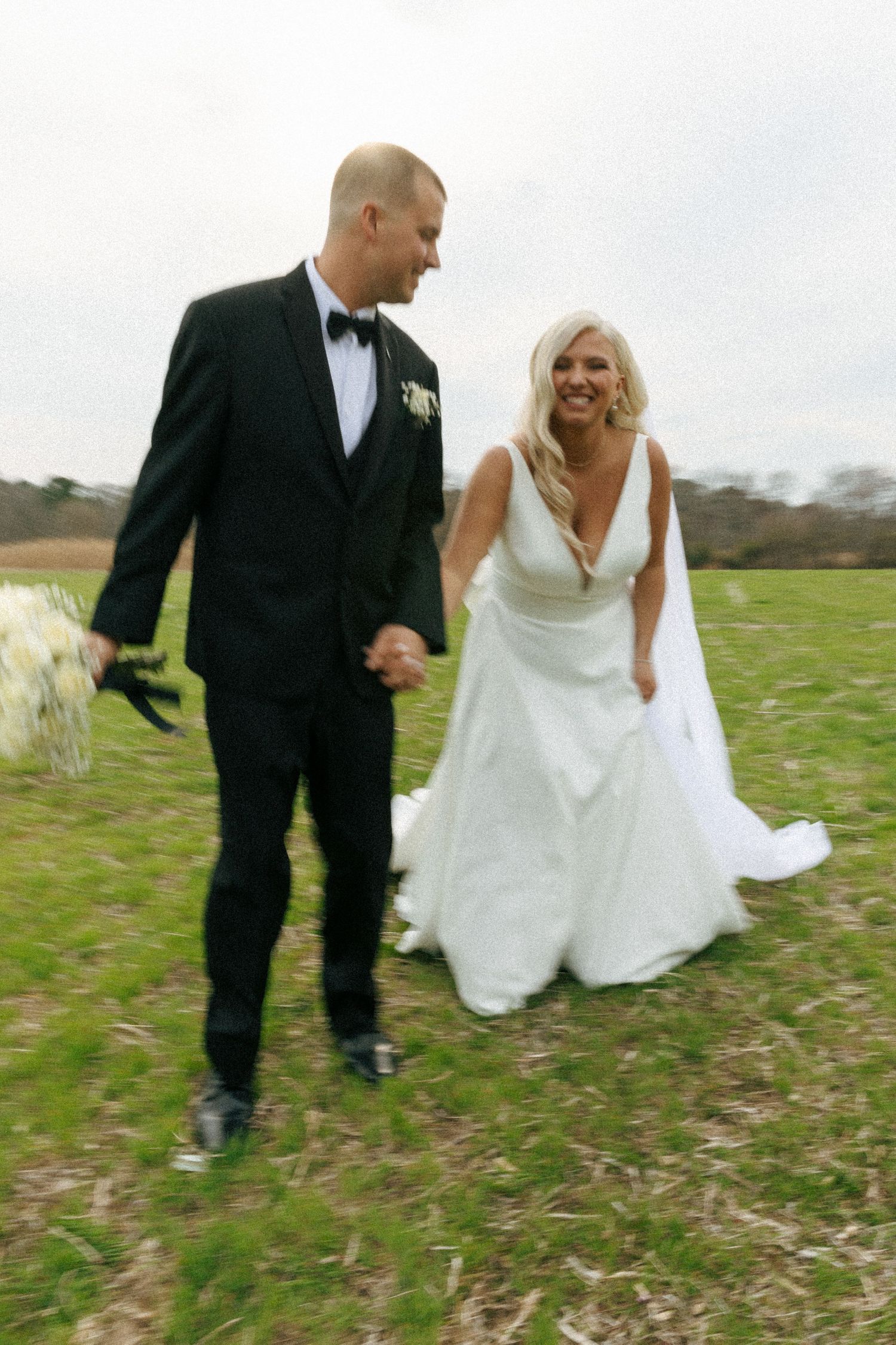 Newlyweds walking together in an open field, with the bride's white dress flowing in the wind.