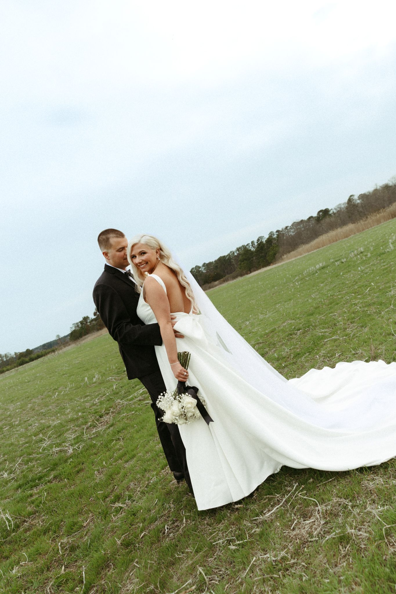 A couple stands in a green field on their wedding day wearing a black tuxedo and flowing white bridal gown with long veil.