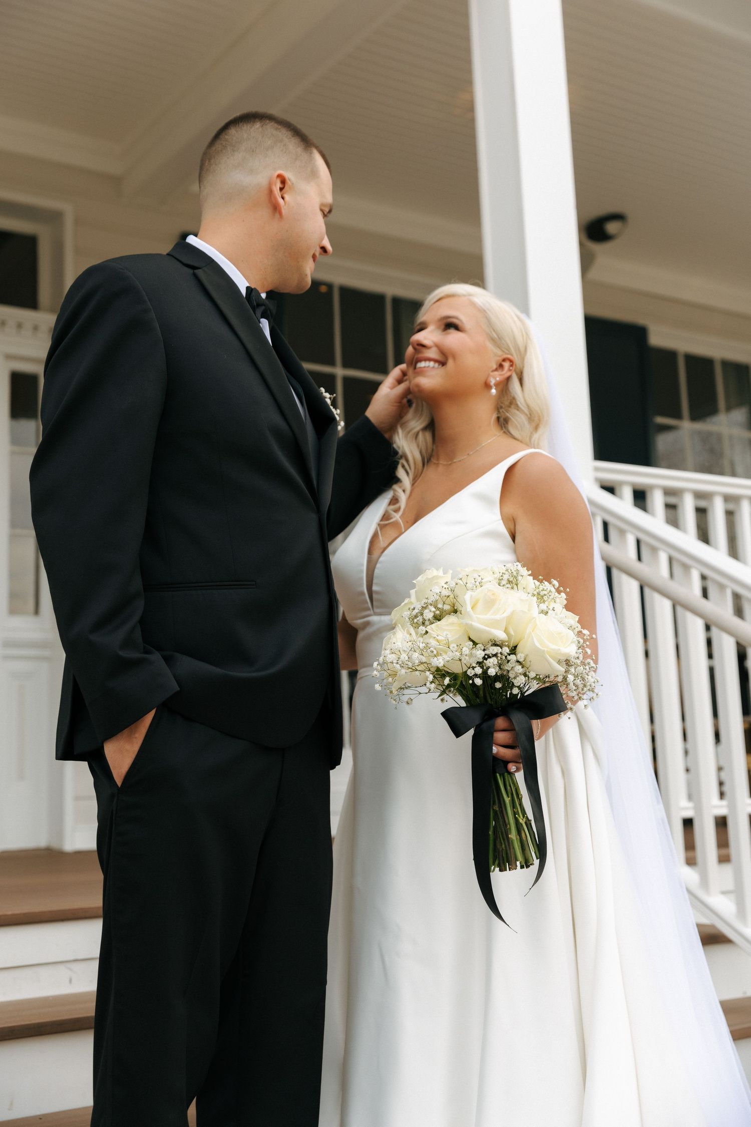A couple share a joyful moment on white porch steps with a bridal bouquet of cream roses.