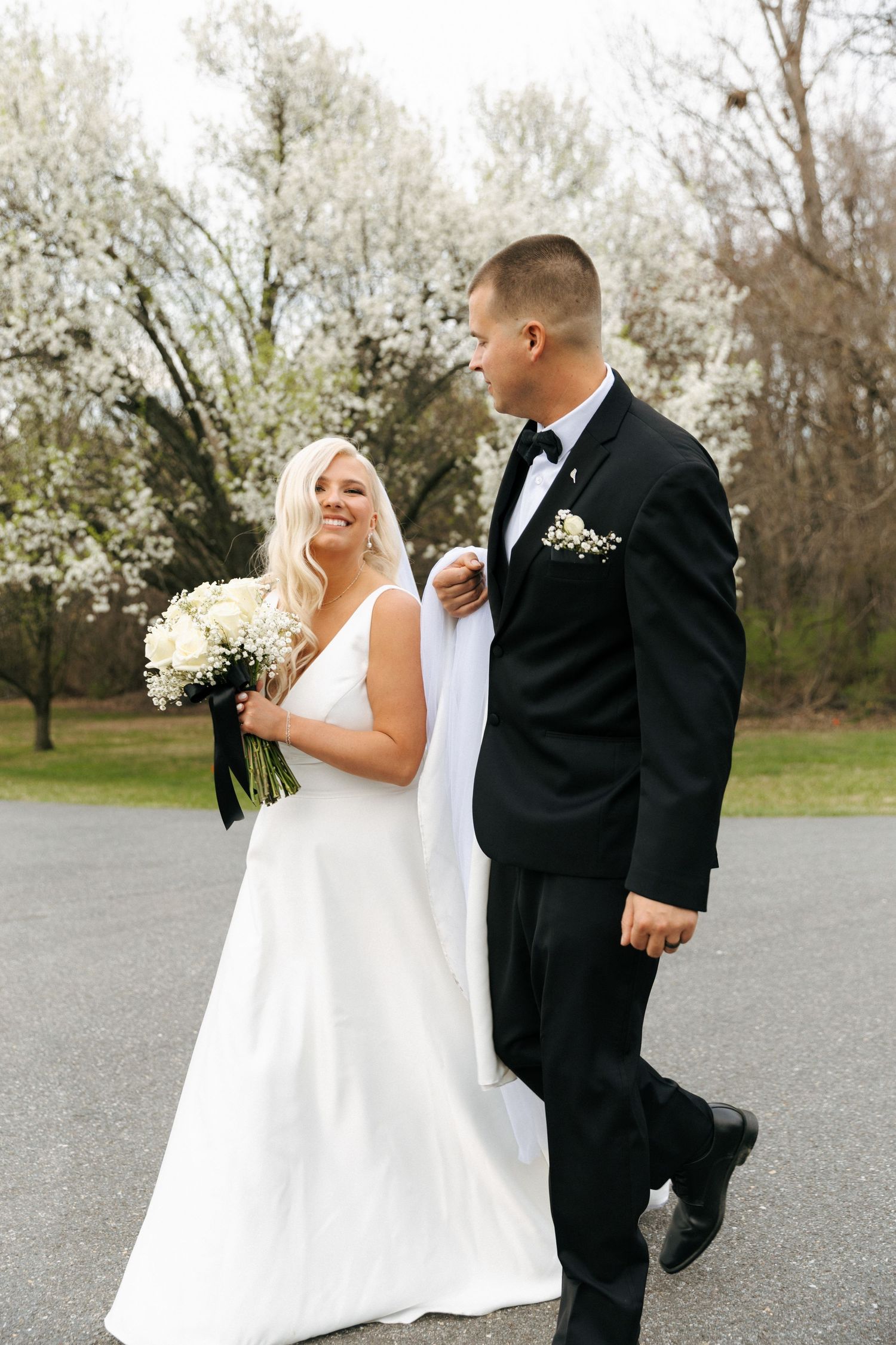 A newlywed couple walking outdoors by blooming white trees with the bride holding a white floral bouquet.