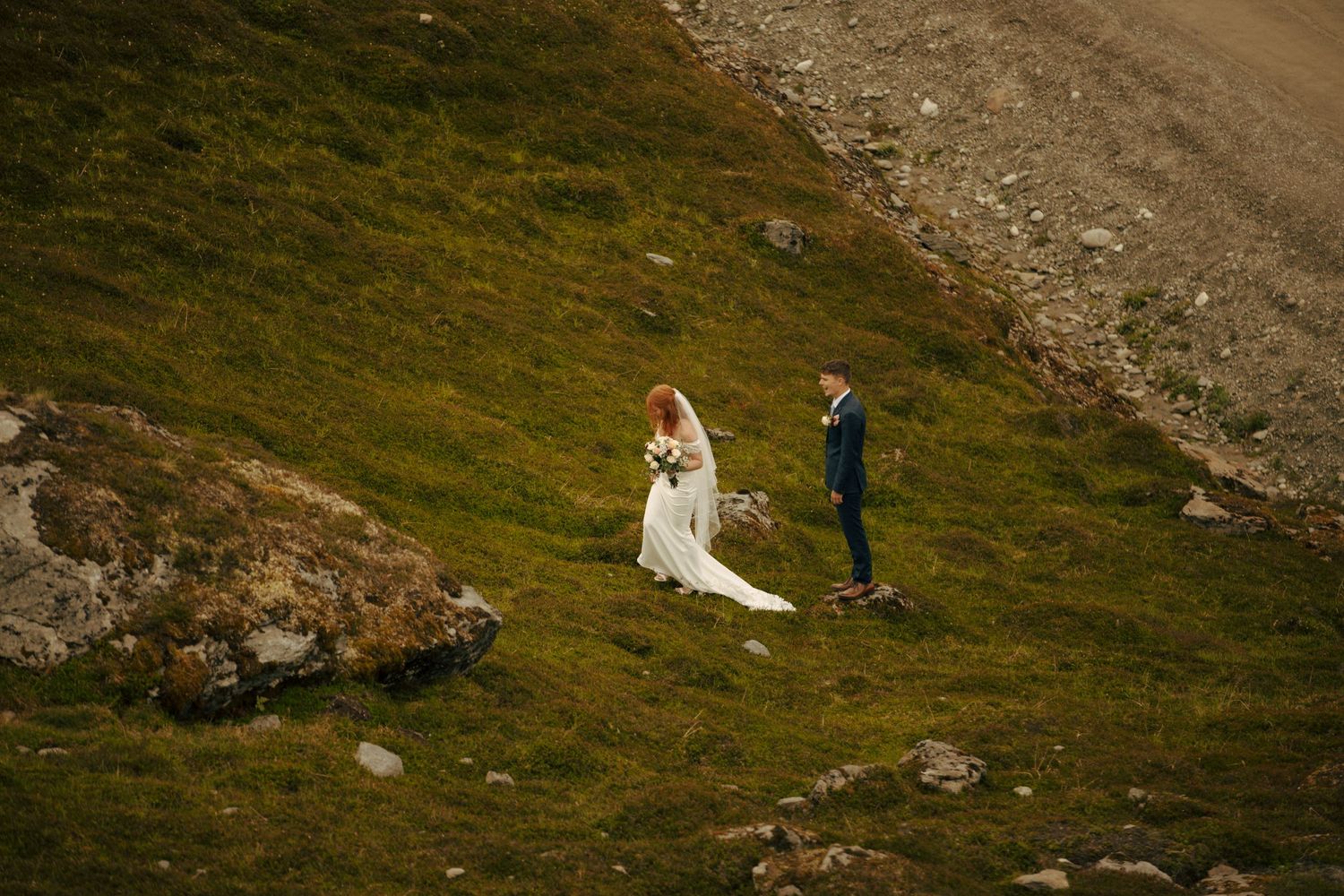 A couple in wedding attire walks along a grassy mountainside slope during an outdoor ceremony.