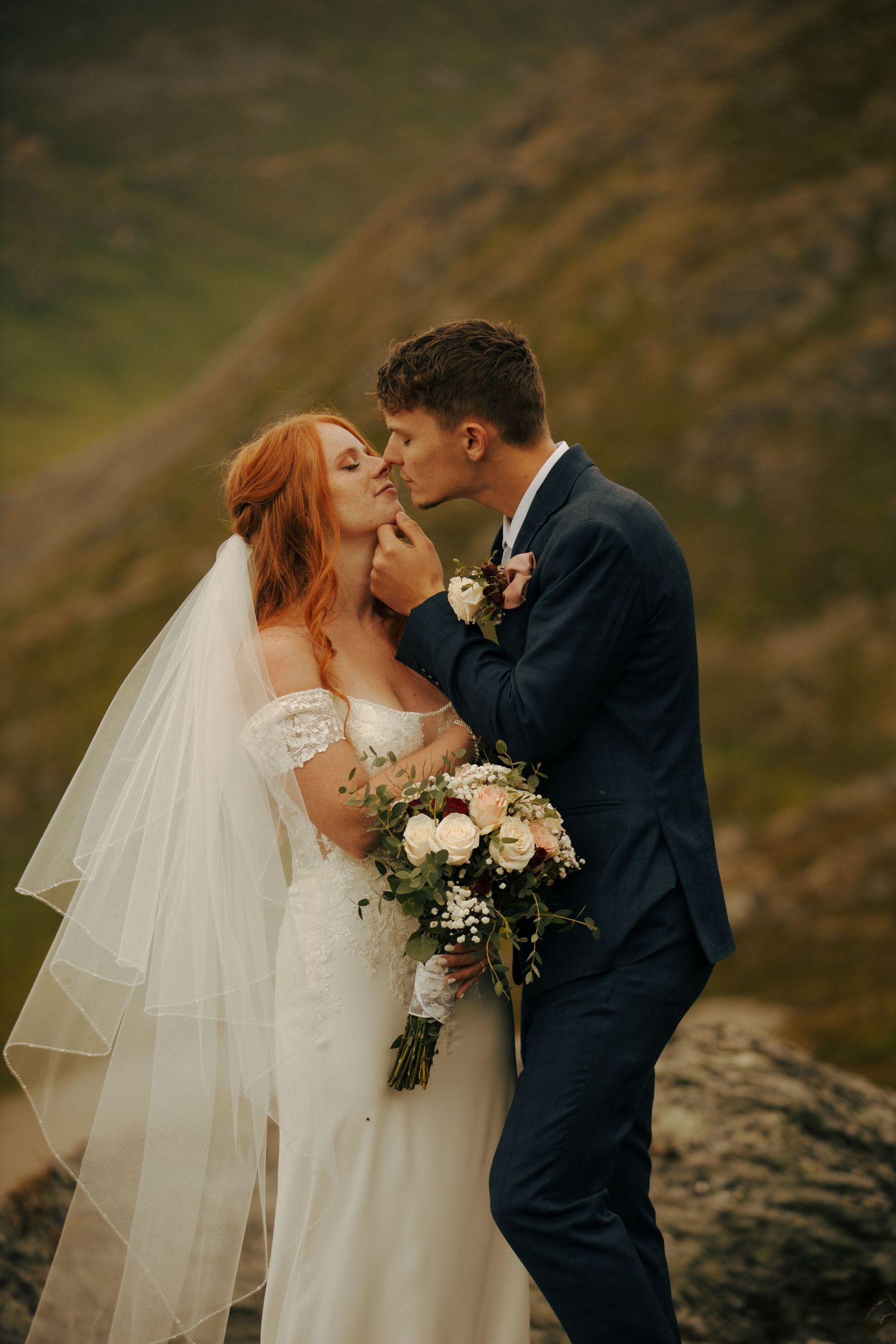 A romantic wedding moment with a couple embracing on a hillside, the bride in white lace holding a neutral bouquet.