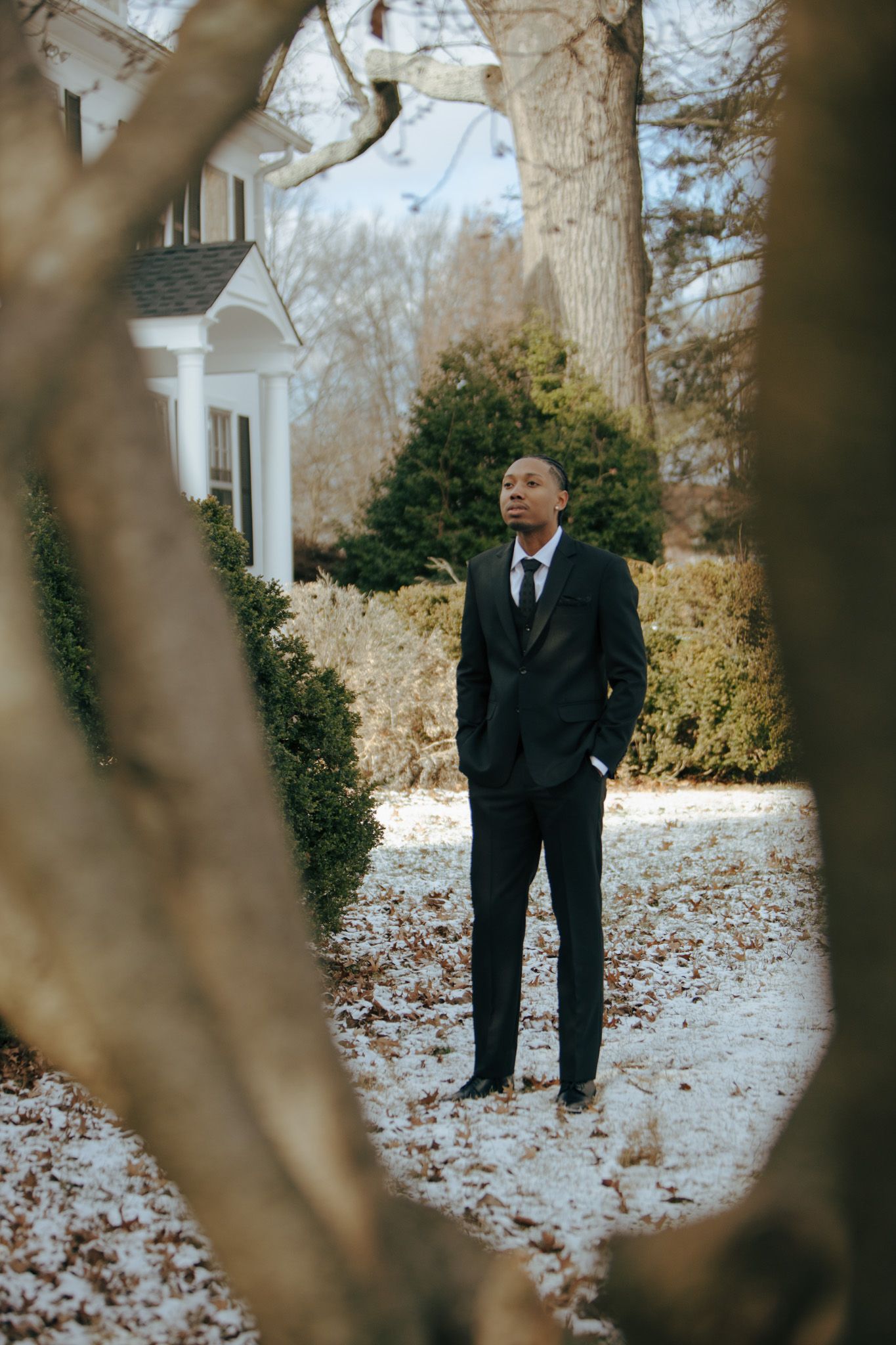 Groom in black suit standing outdoors in winter garden setting.