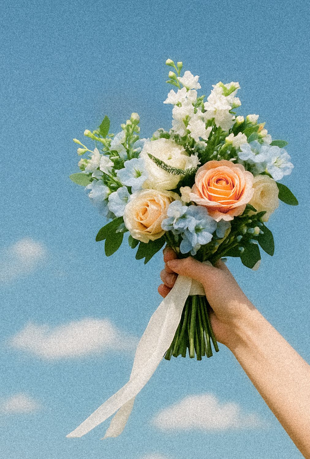 A hand holds up a romantic bouquet of peach roses and white delphinium flowers against a bright blue sky with clouds.