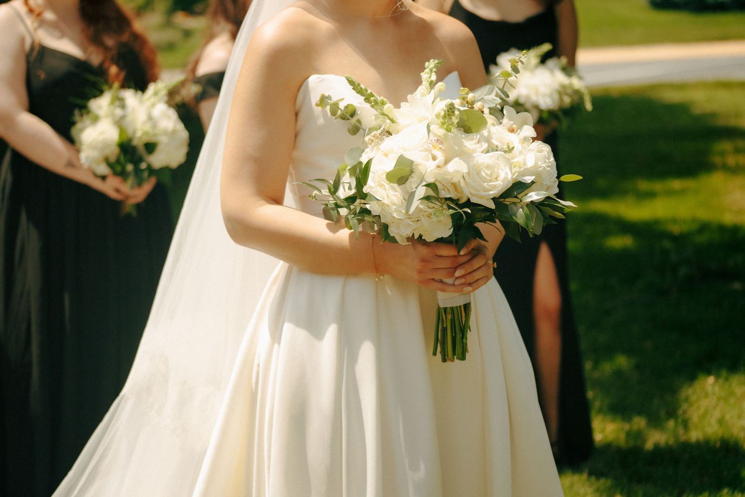 Sunlit wedding scene showing white bridal gown and bouquet with bridesmaids in dark dresses.