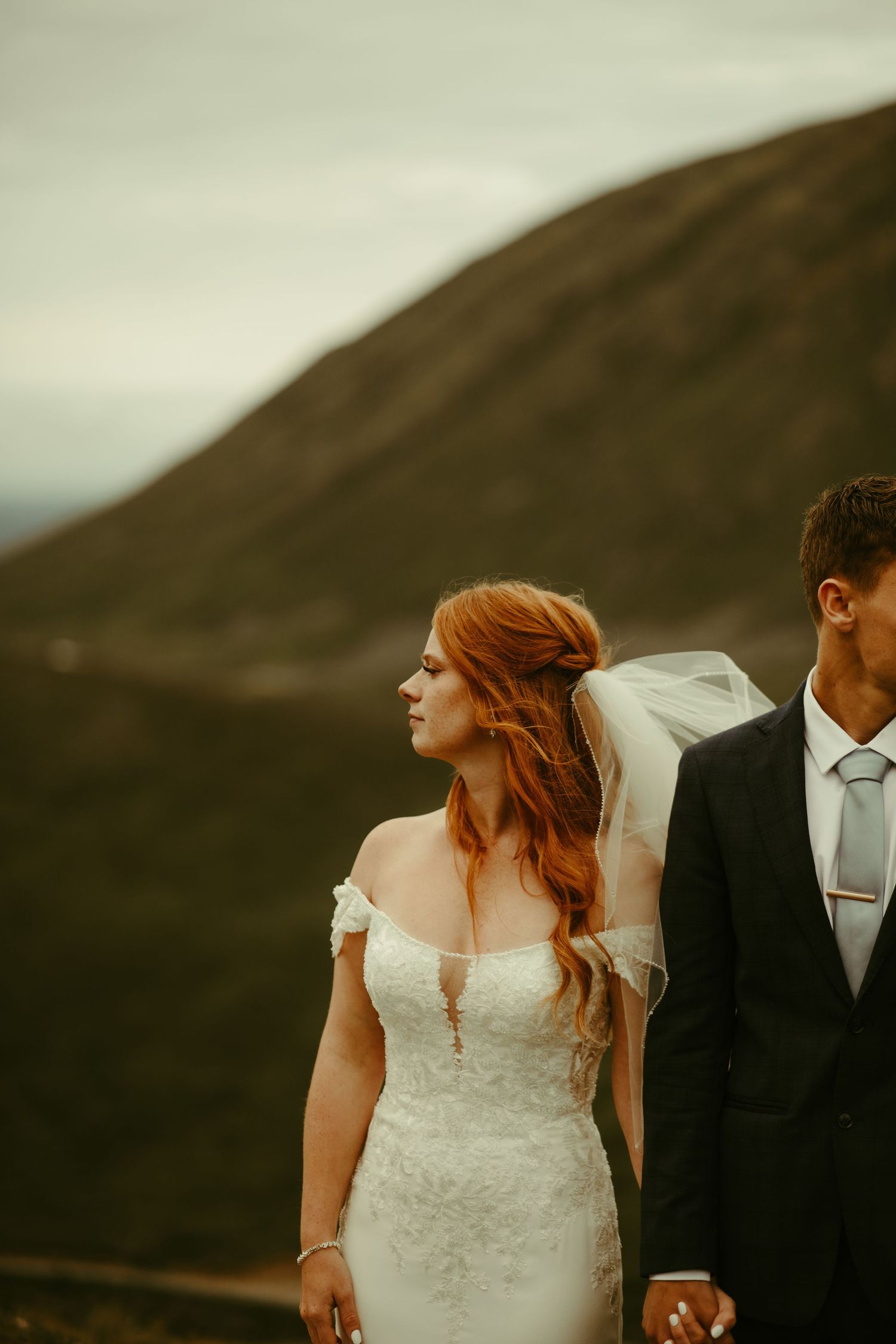 A wedding couple stands against misty mountain backdrop with bride in lace dress and groom in dark suit.