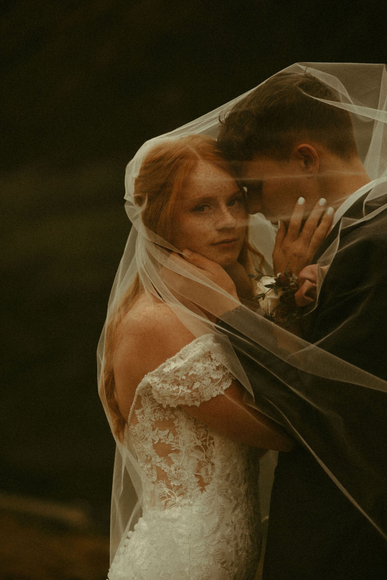 An ethereal wedding portrait captures a tender moment between newlyweds beneath a delicate veil at dusk.