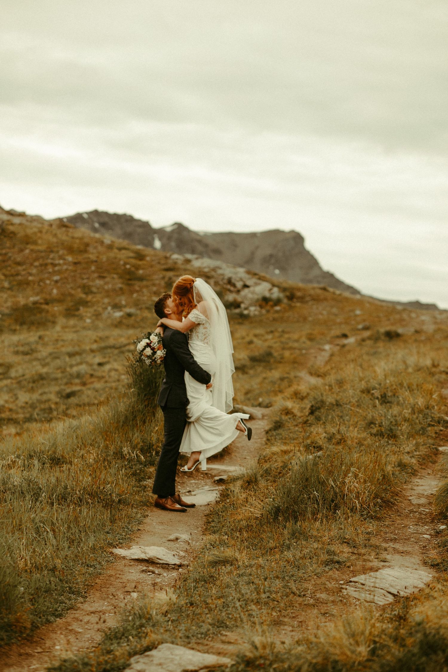 A couple embraces on a mountain trail with golden grass and rocky peaks in the background under an overcast sky.