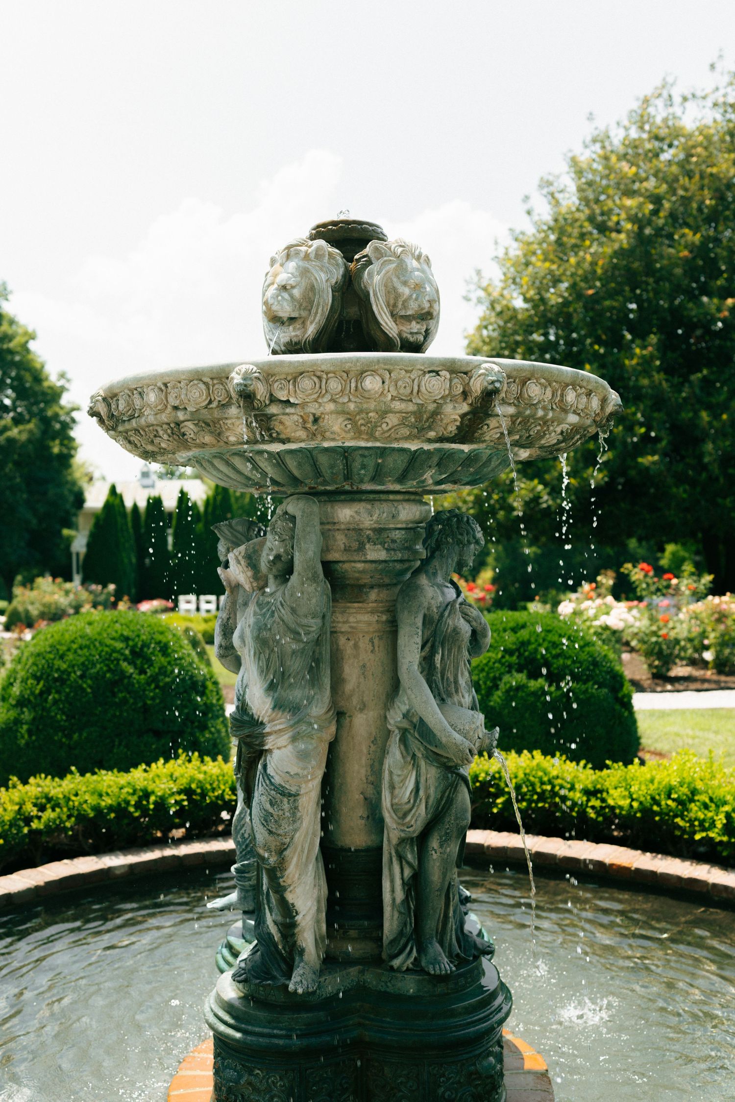 Ornate stone fountain with cherub sculptures spraying water in a formal garden setting with manicured hedges and trees.
