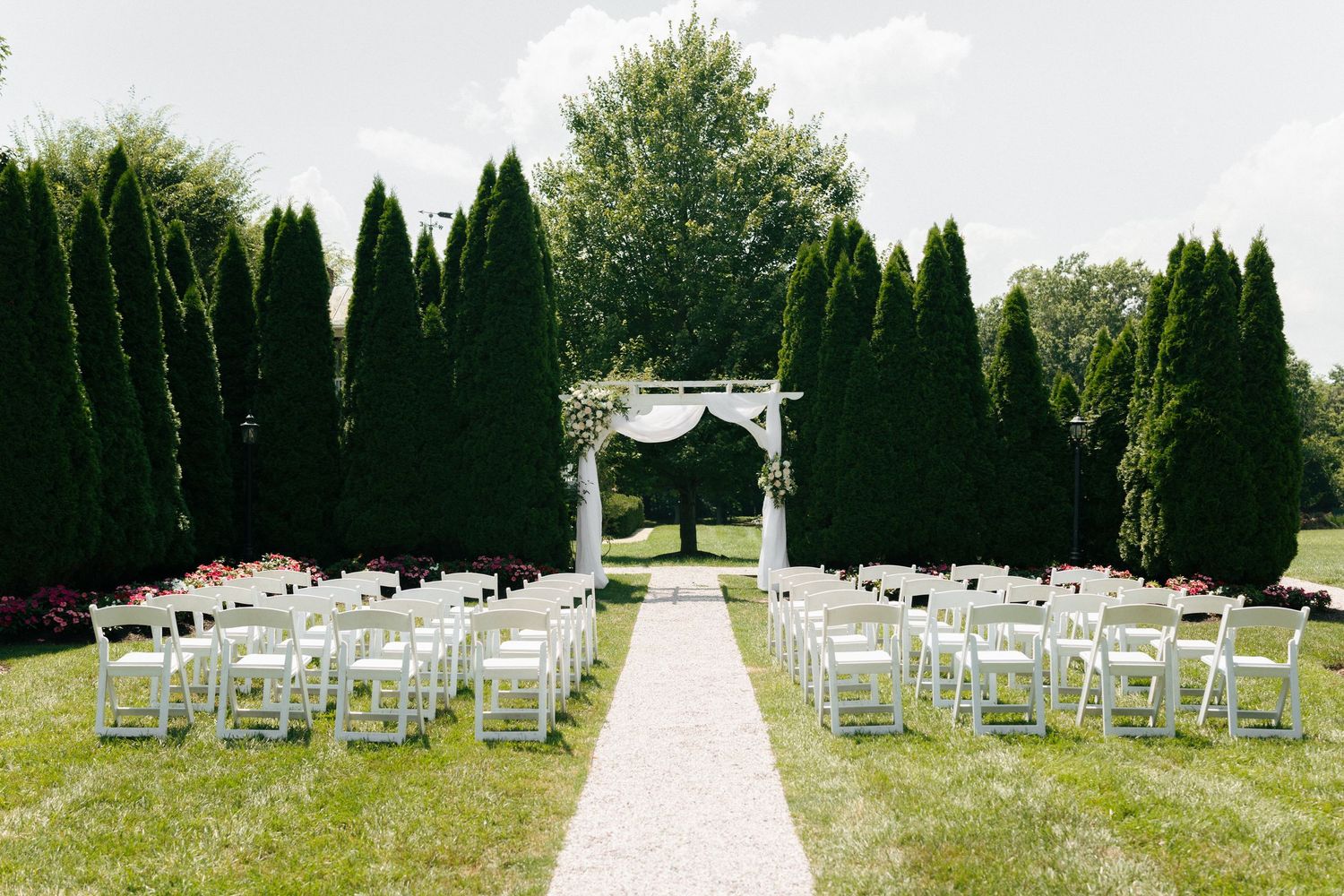 White chairs arranged in rows for an outdoor wedding ceremony with tall trees and an elegant white arch.