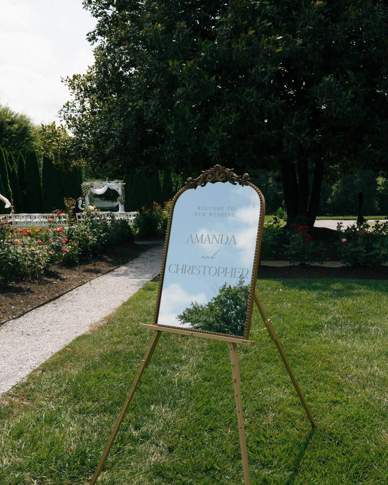 An ornate gold-framed welcome sign on an easel stands along a garden pathway with trees in the background.