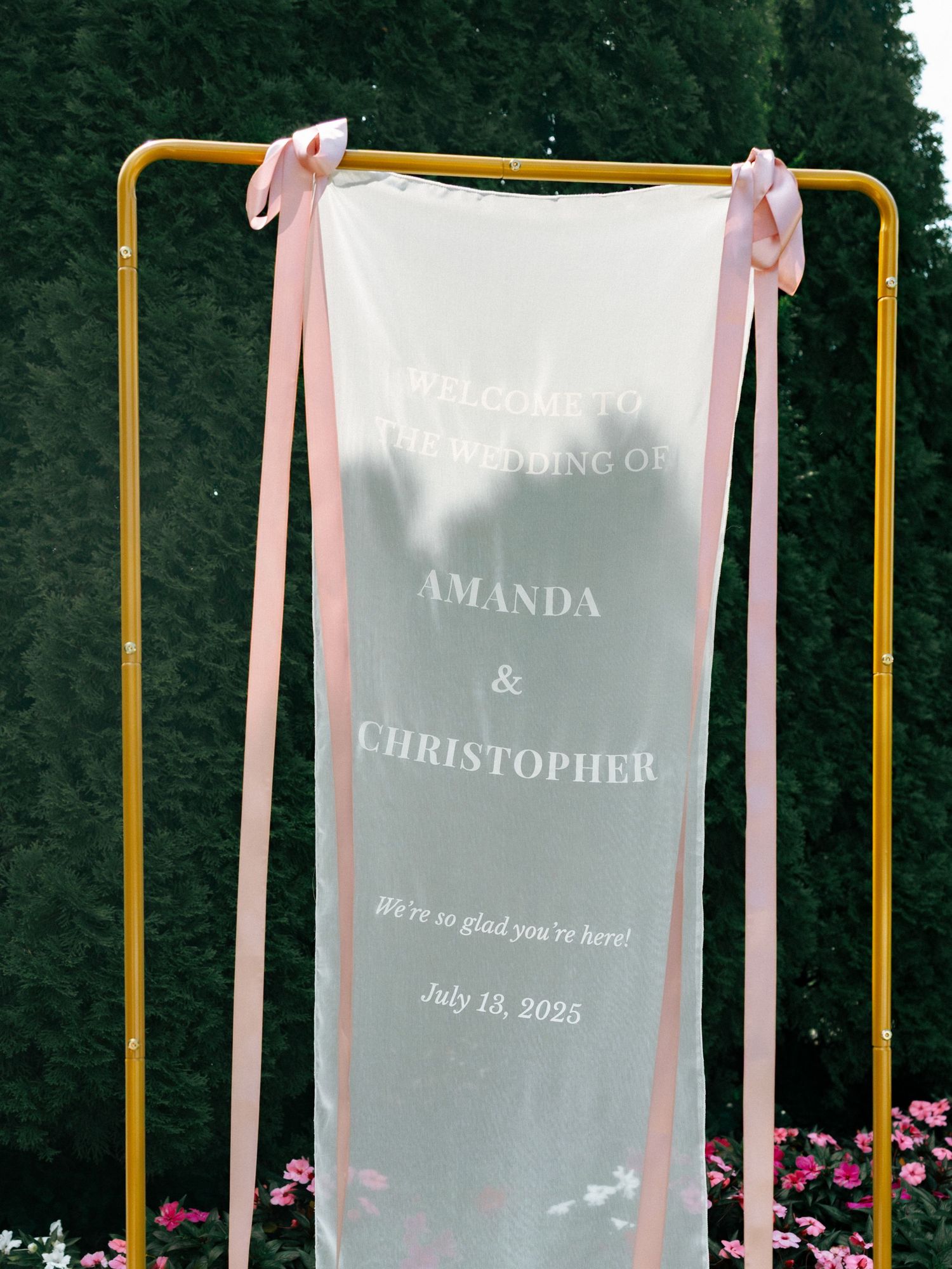 A white wedding sign with pink ribbons hangs on a yellow metal frame outdoors.