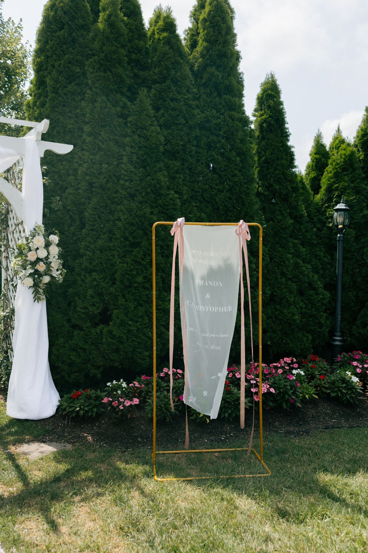 White dress displayed on a copper clothing rack against evergreen trees and pink flowers in a garden setting.